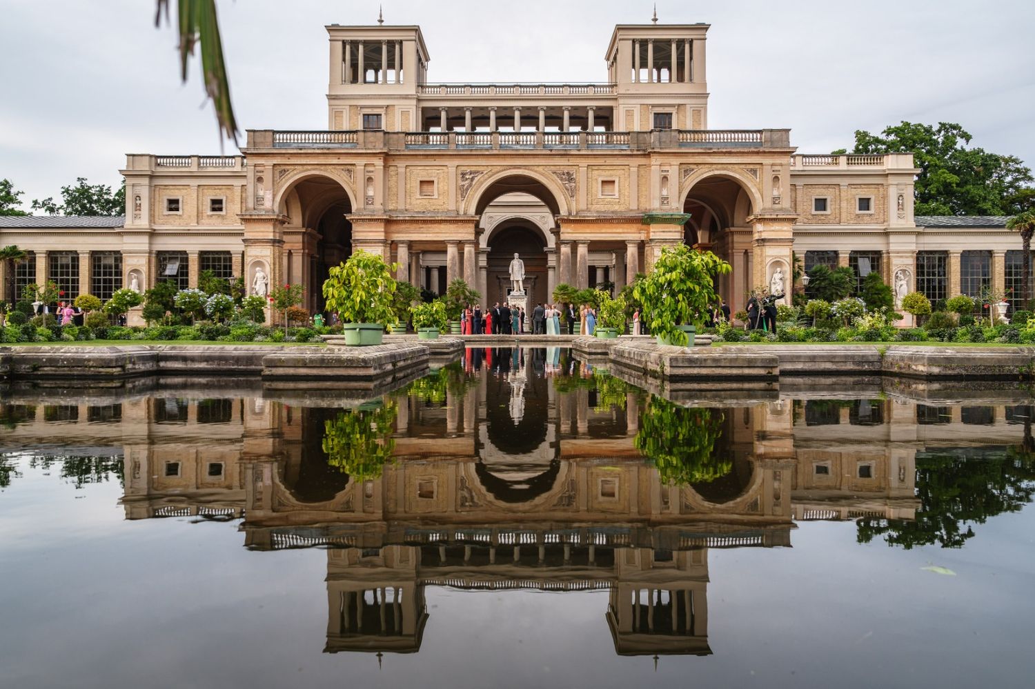 Historic palace building with ornate architecture reflects perfectly in still water surrounded by lush gardens and trees.