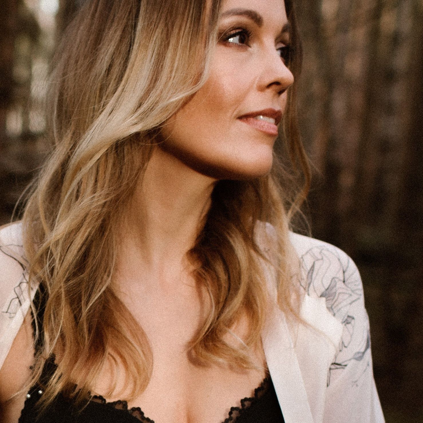 Soft portrait in warm tones showing blonde wavy hair and black lace top against blurred forest background.