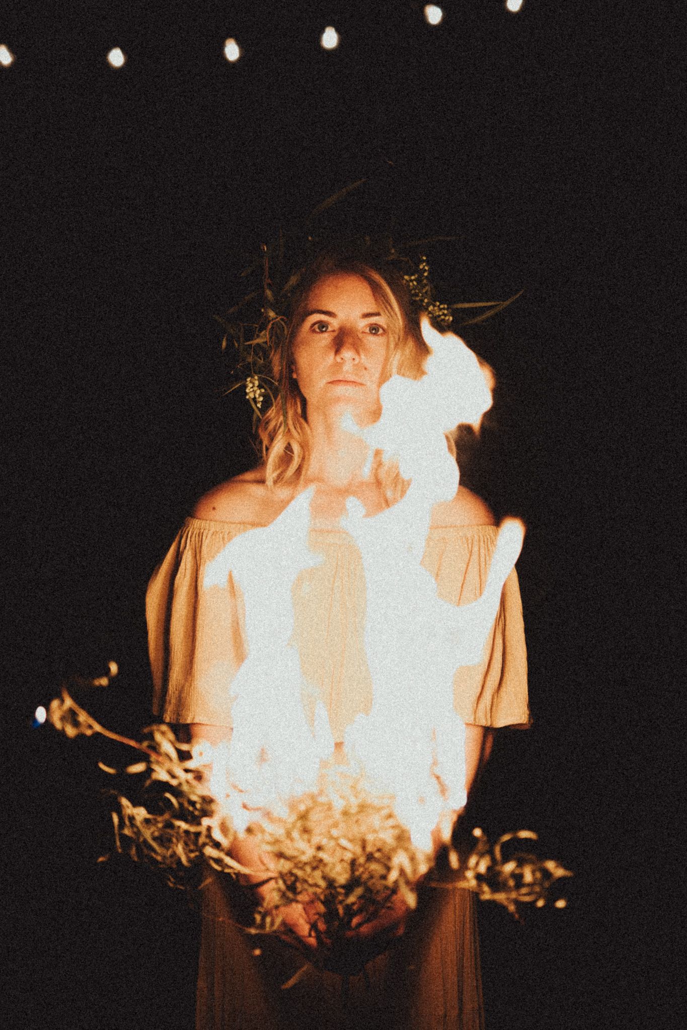 Moody nighttime portrait with glowing fire flames illuminating a figure in a beige top against a dark background.
