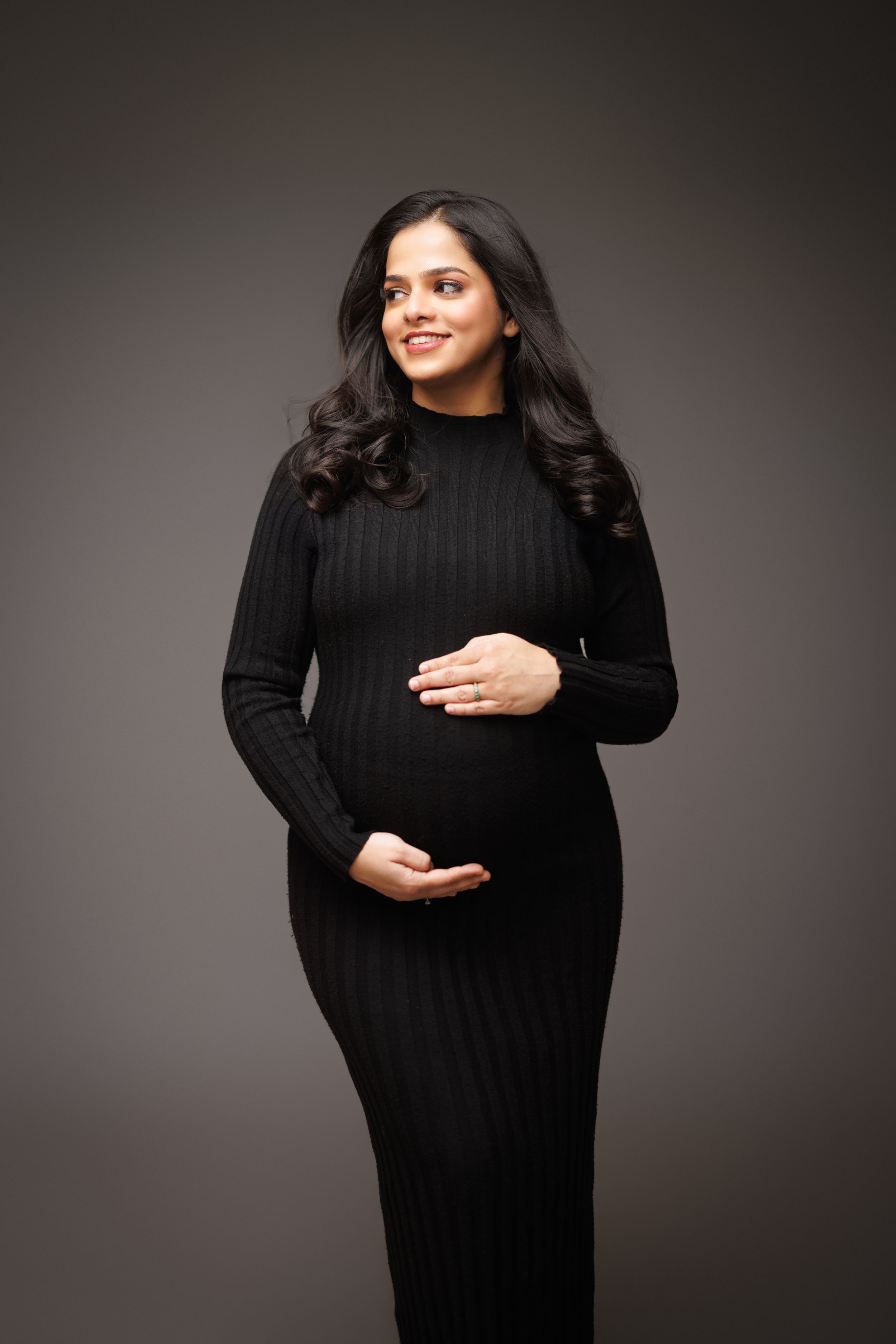 Studio maternity portrait in fitted black sweater dress against grey backdrop.