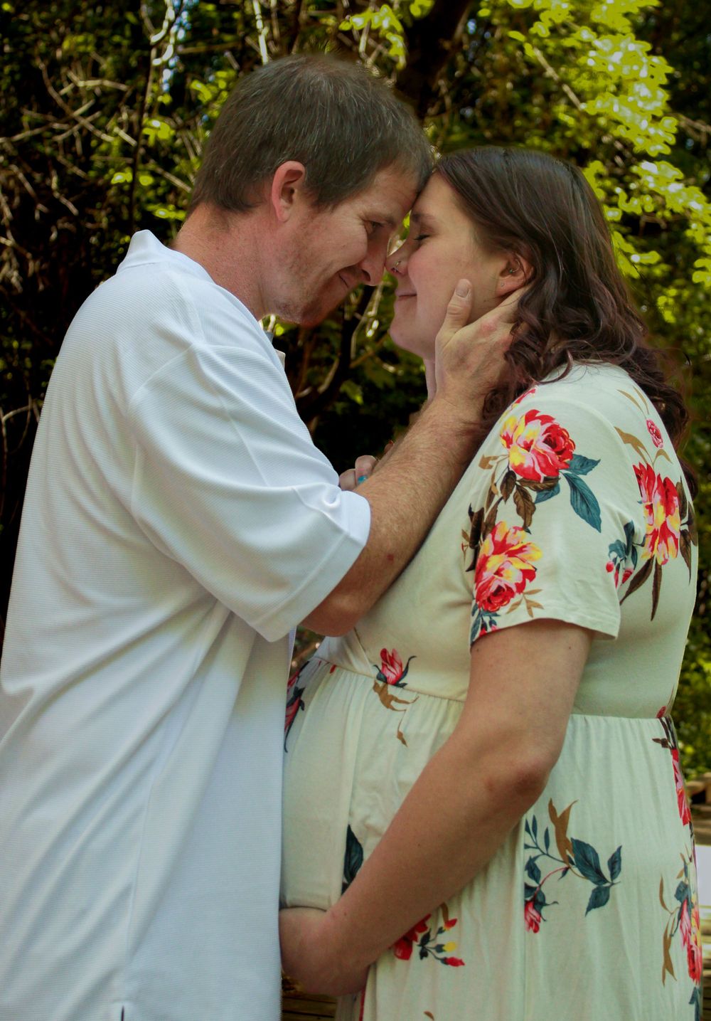 A couple shares an intimate moment in a garden setting, with her wearing a light floral dress and him in white.