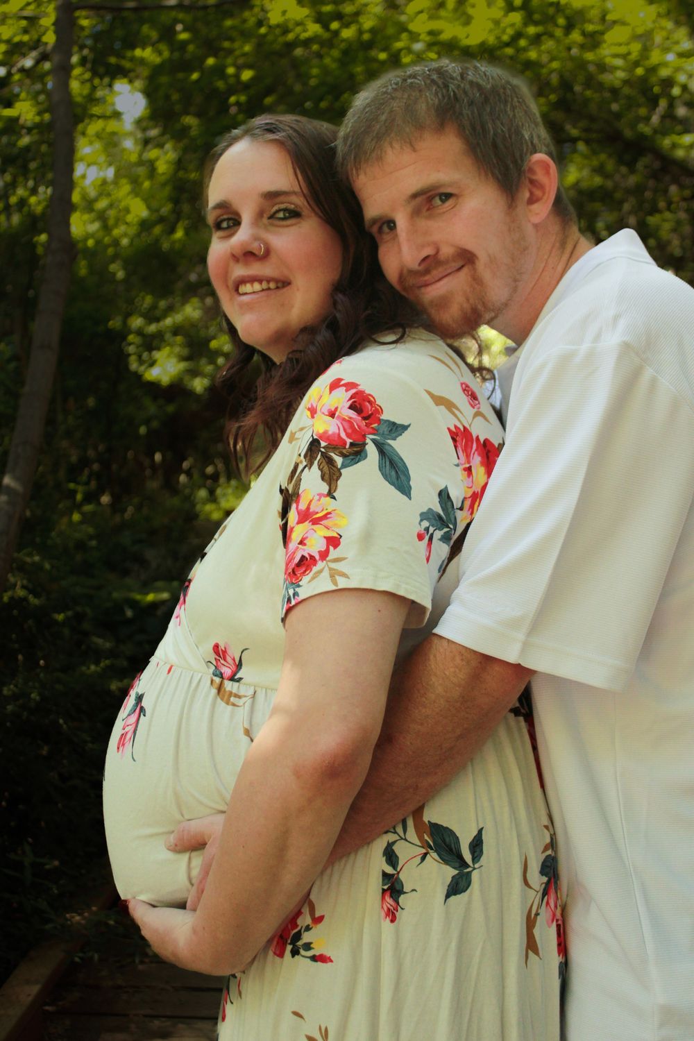 Romantic maternity photo of couple embracing outdoors with floral dress against forest backdrop.