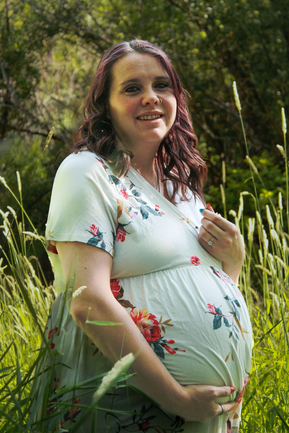 A smiling person in a floral dress poses playfully against a backdrop of tall grass and greenery.