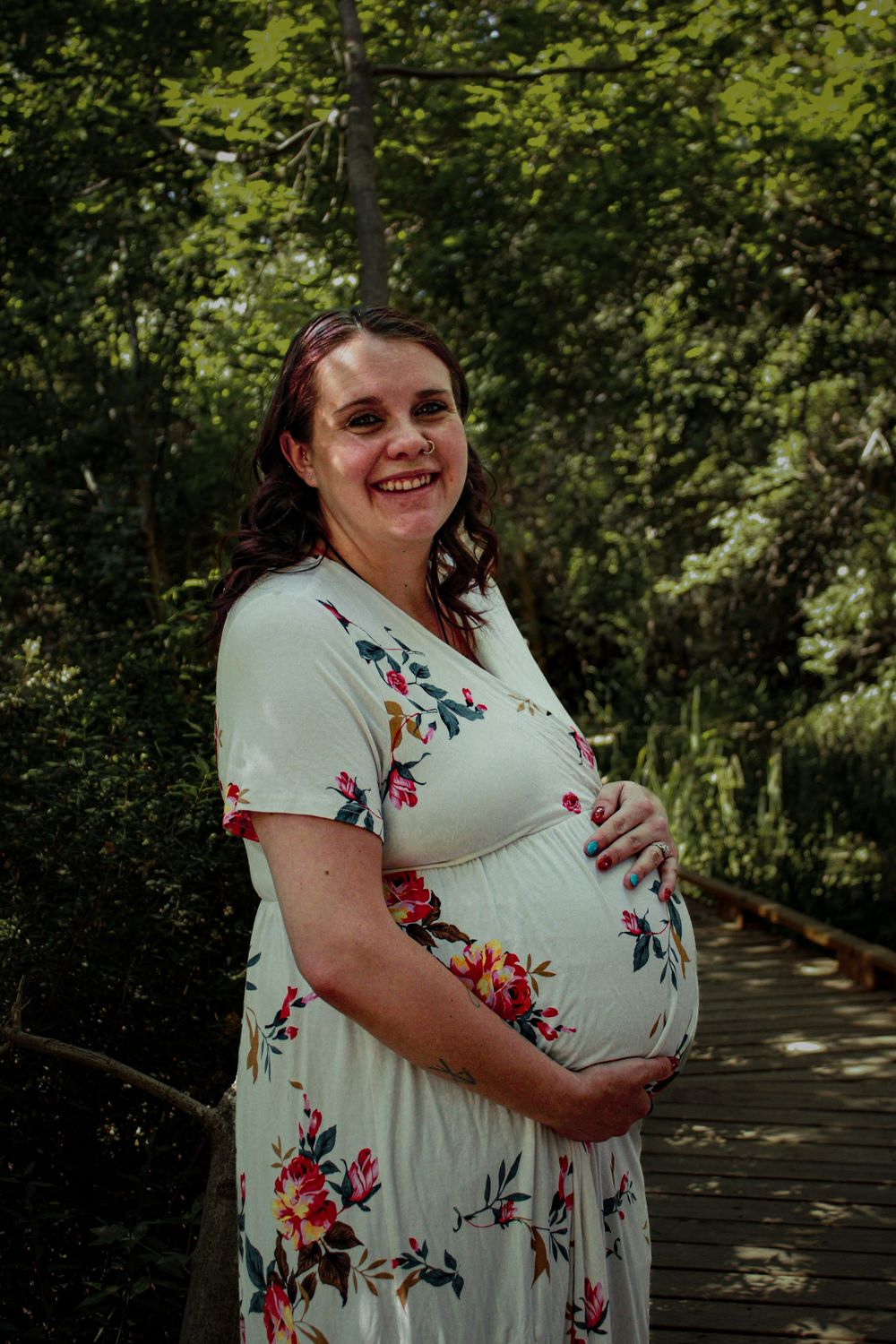 Expectant mother in floral dress smiles while posing on wooden walkway surrounded by greenery.