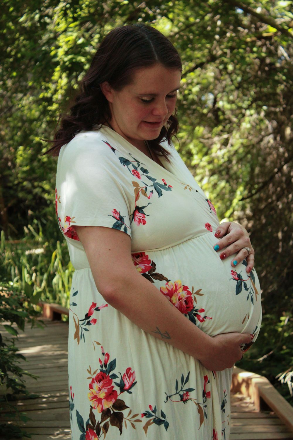 Expectant mother in a floral white dress cradles her belly while standing on a wooden walkway surrounded by greenery.