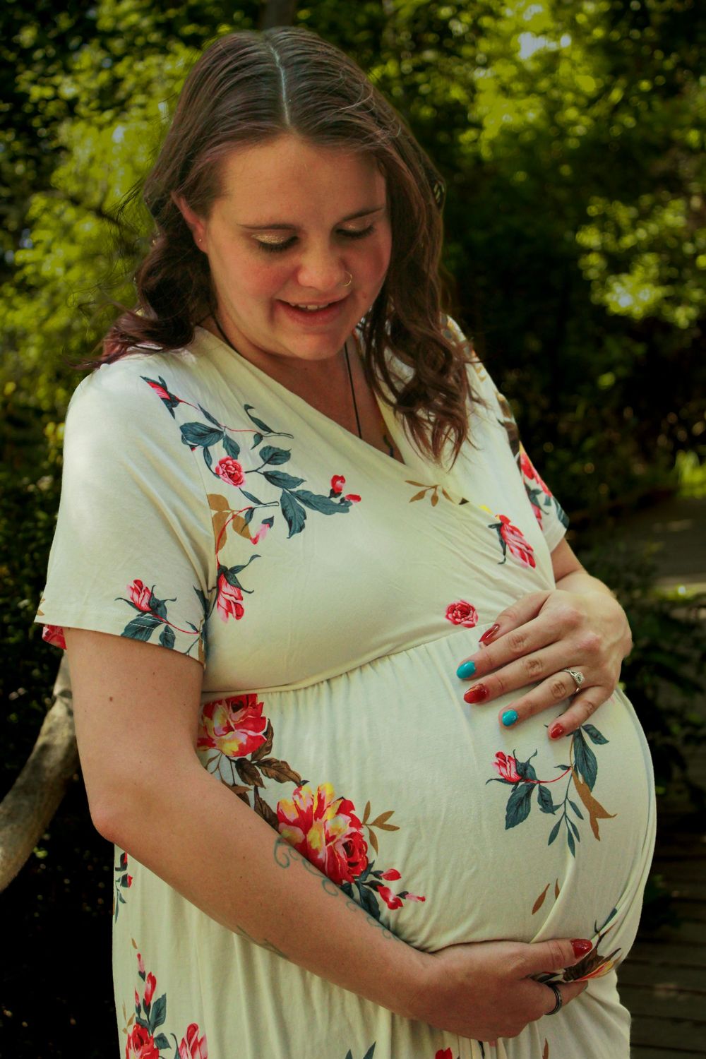 A pregnant woman in a floral dress smiles while holding her baby bump outdoors in a garden setting.