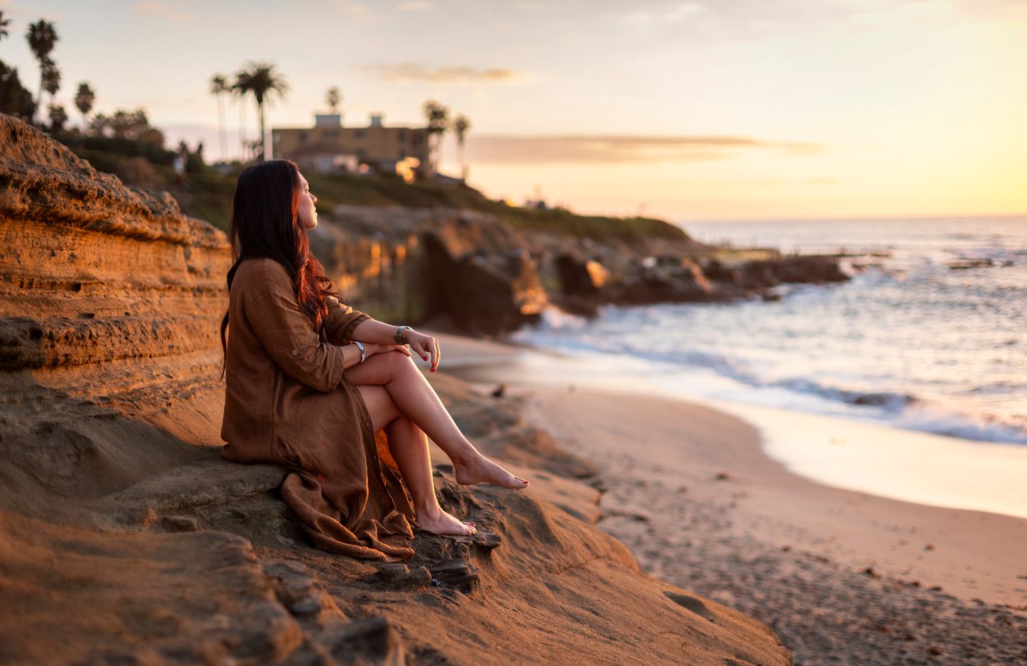 Woman sitting on sandy cliff overlooking ocean at golden sunset.