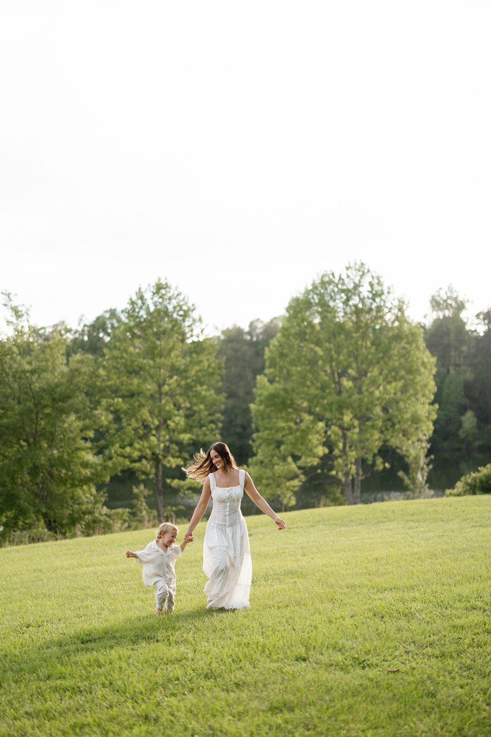 A mother and small child run together across a sunny green hillside surrounded by trees.