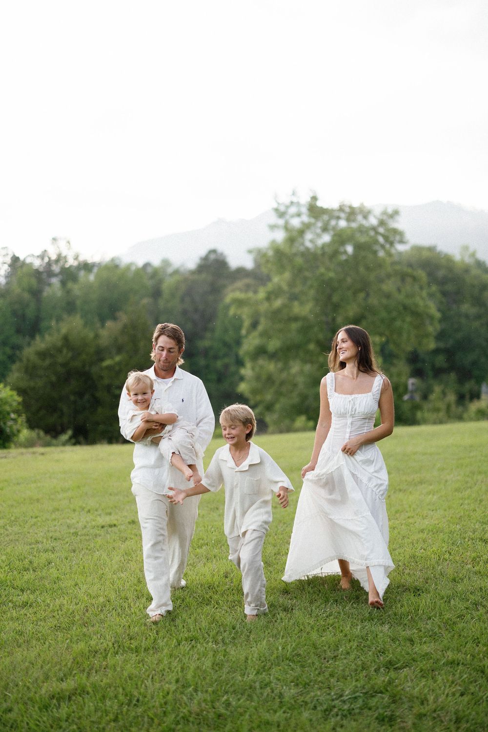 A joyful family wearing white outfits runs and plays together in a lush green field on a sunny day.