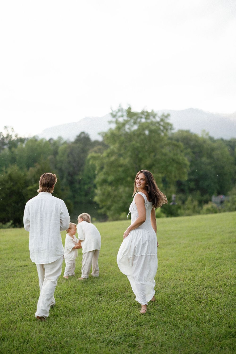 A family in white clothing walks together across a serene grassy hillside on a summer day with mountains in the distance.
