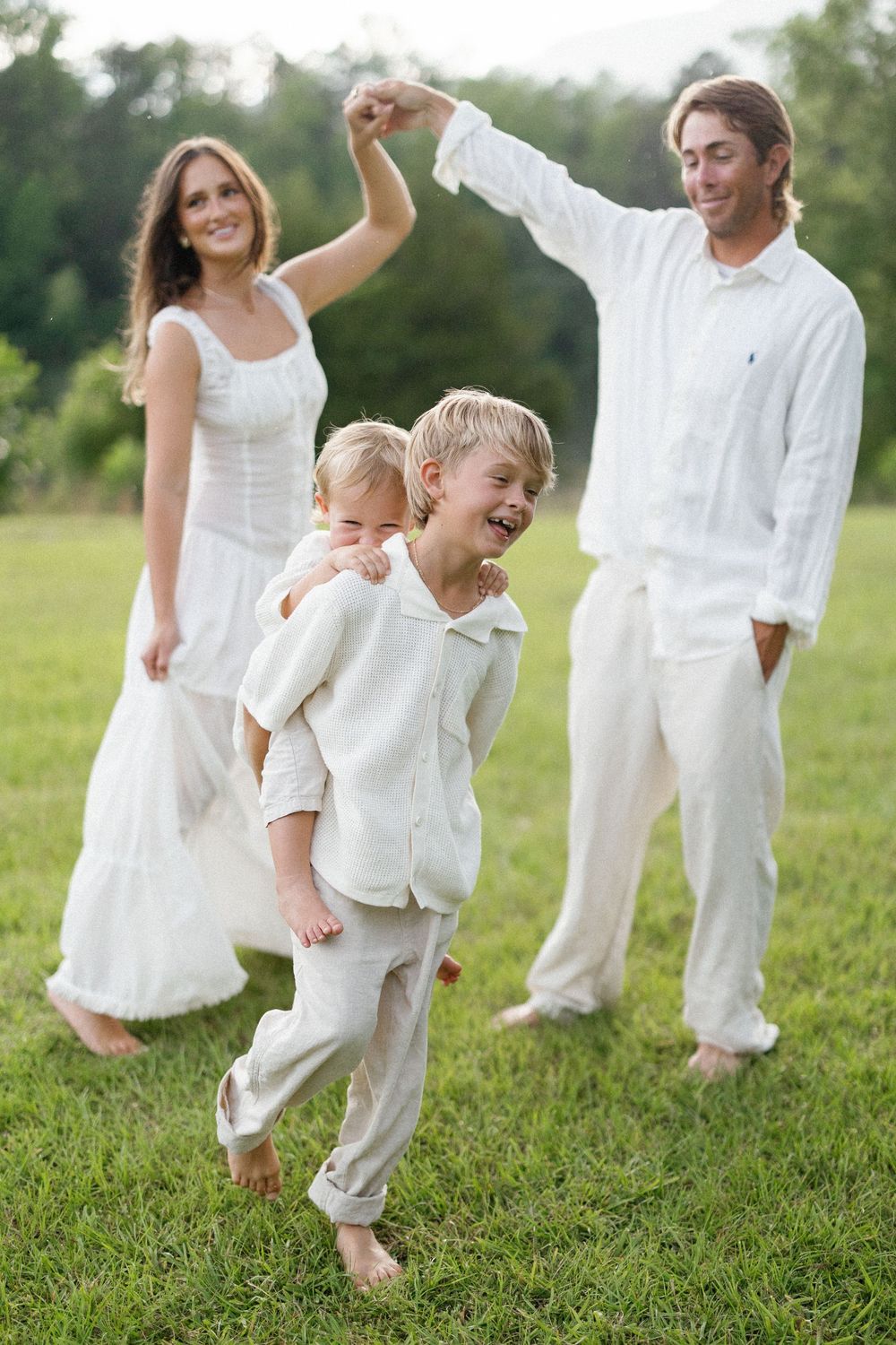 A family dressed in white dances together on grass, with a young child laughing and playing in the foreground.