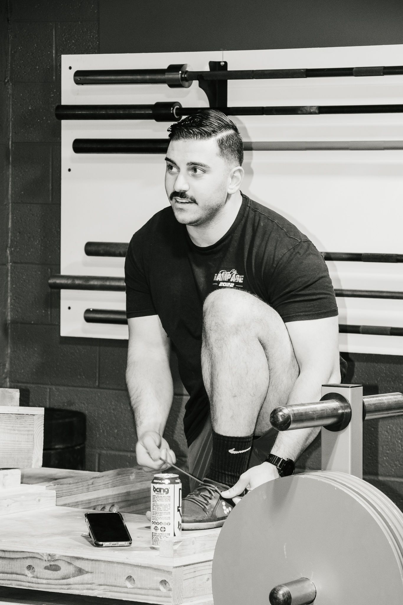 Man in gym clothes kneels to tie his shoe beside weights, with a Bang energy drink and phone on a wooden platform.