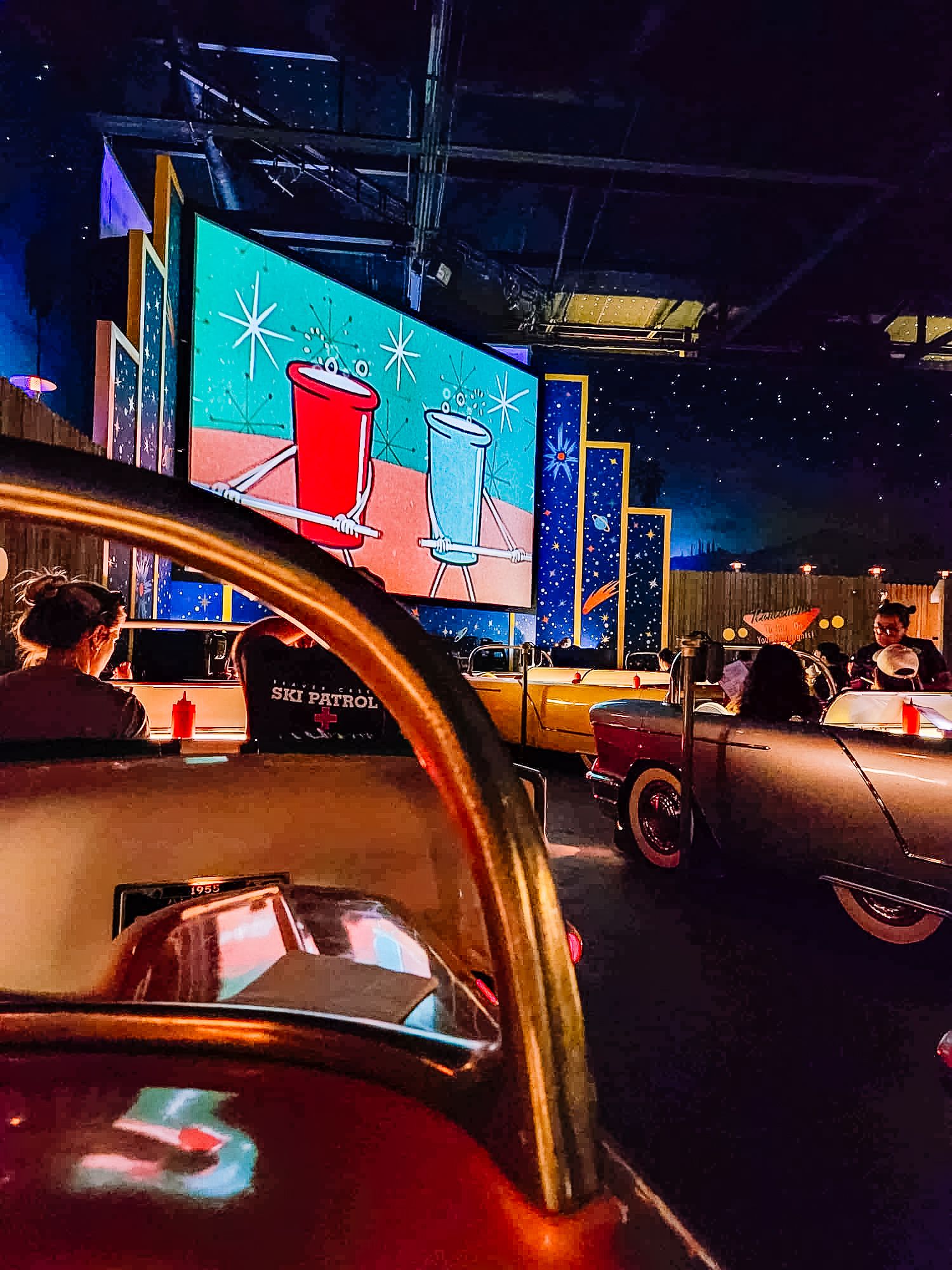 Drive-in theater style seating with vintage cars facing large projection screen.