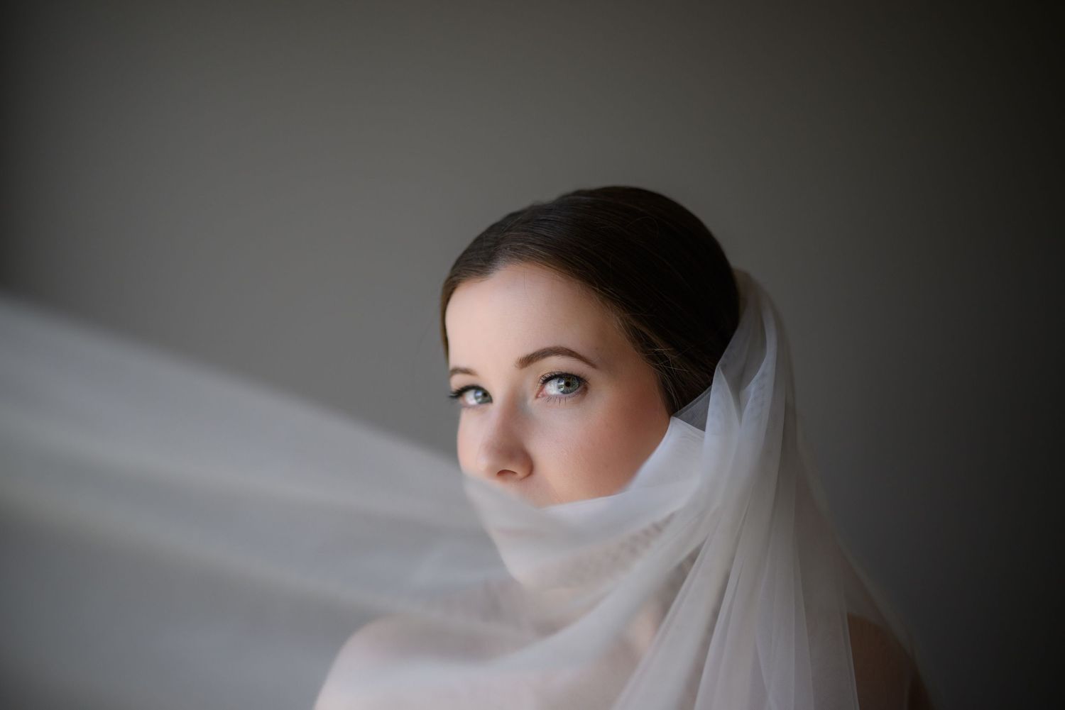 A dreamy portrait of bride wearing an ethereal white veil, gazing upward with a thoughtful expression in soft lighting.
