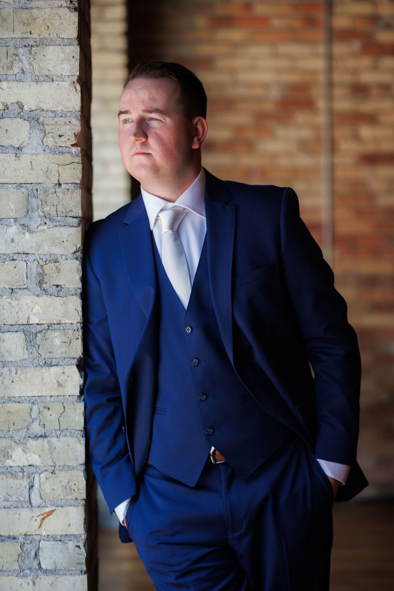 A groom in a tailored navy blue three-piece suit and white tie poses against a brick wall.