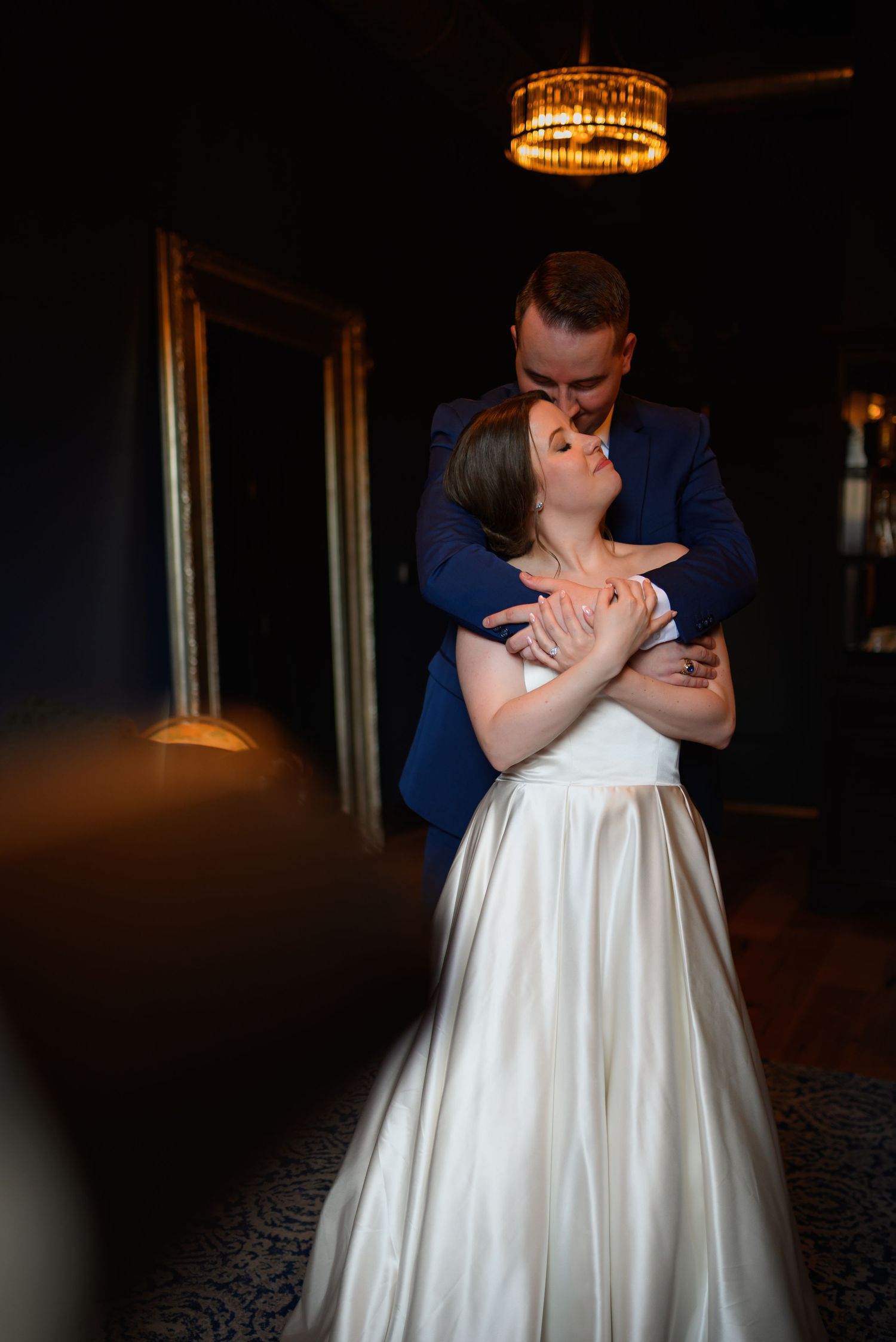 A couple embraces romantically on their wedding day with the bride in a white gown and groom in navy suit under warm lighting.