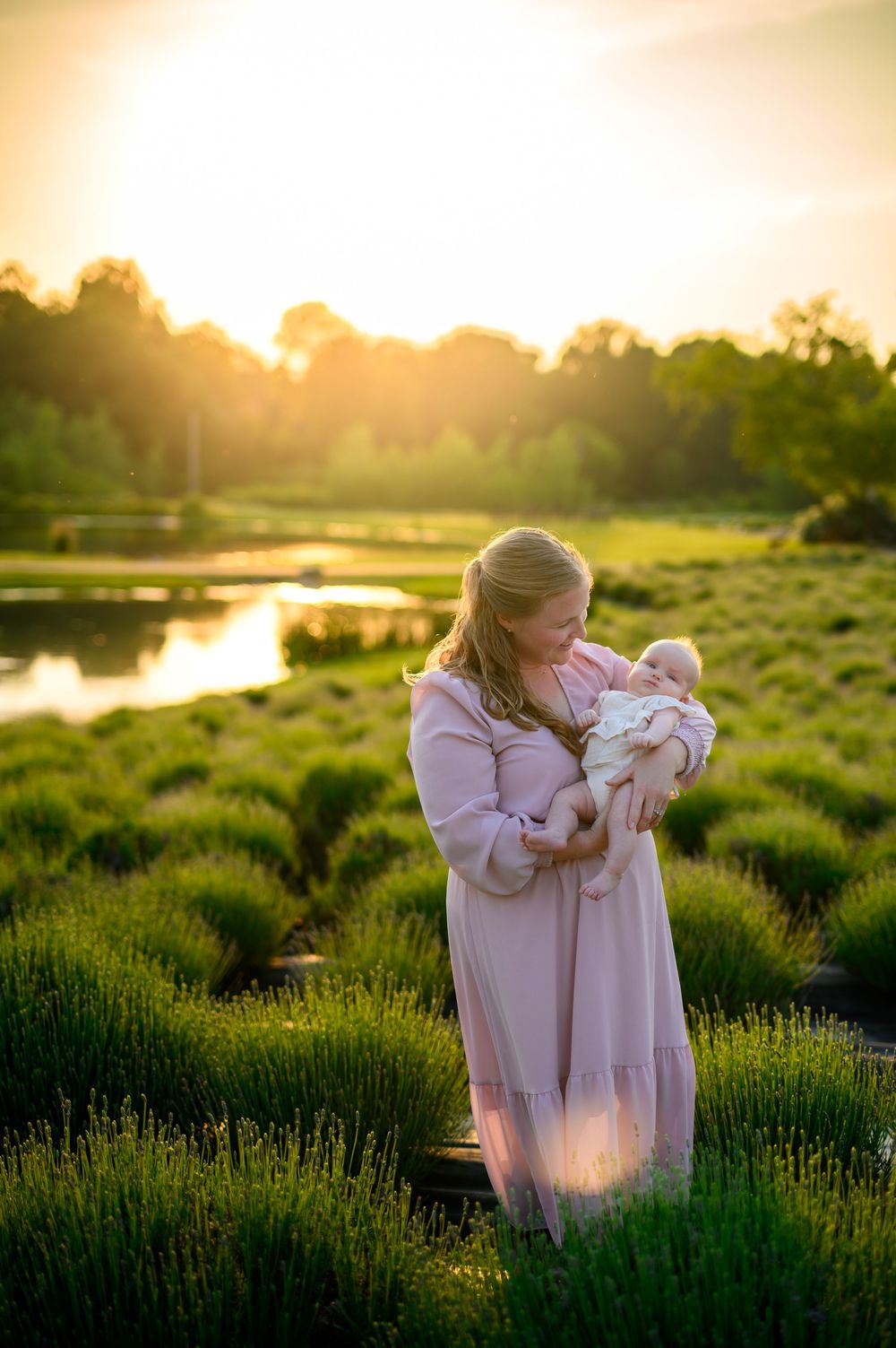 A woman in a pink sweater holding a small baby at sunset in a lavender field with golden light streaming behind them.