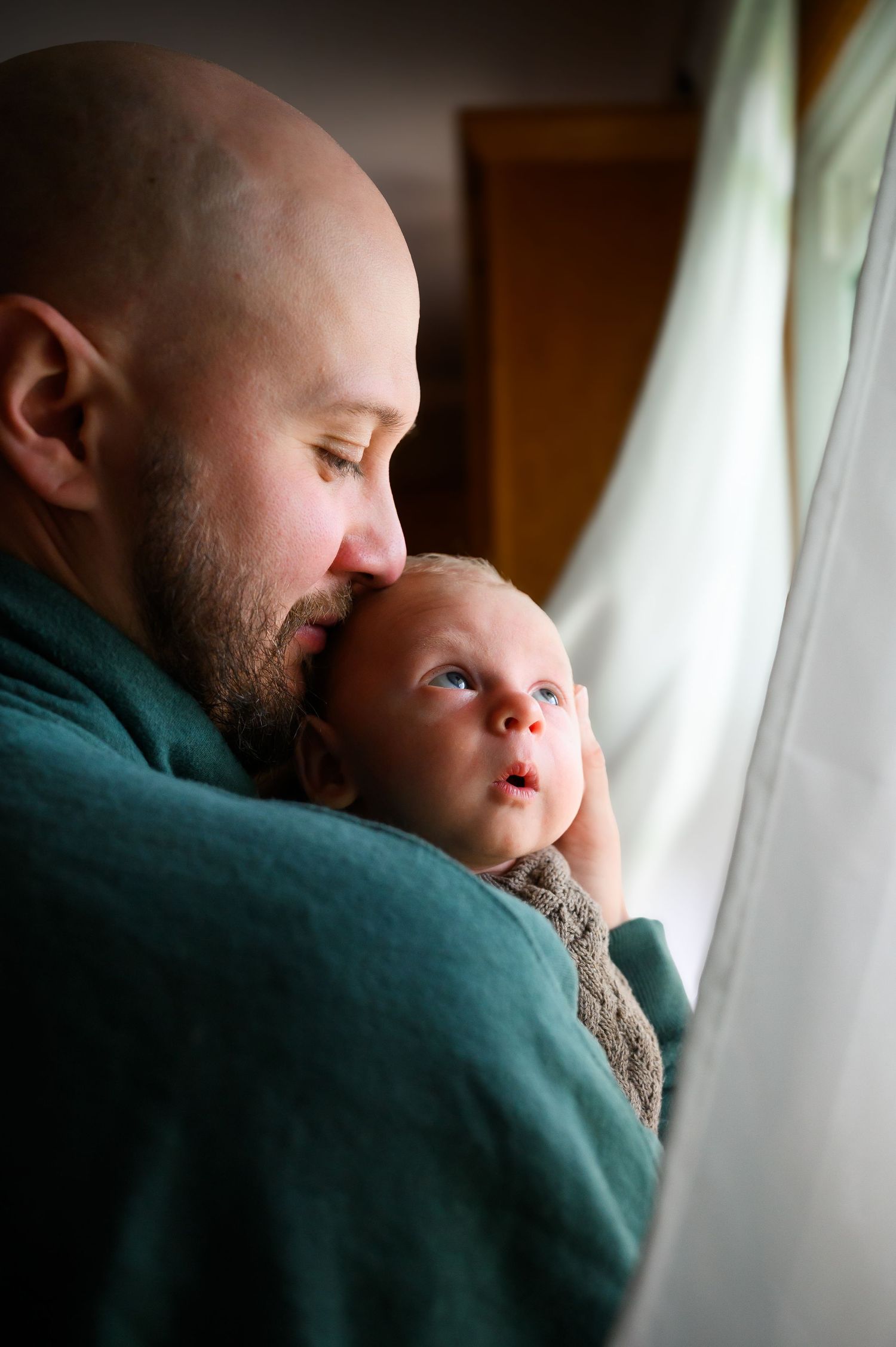 Bald bearded father tenderly holding newborn baby near window in soft natural light.