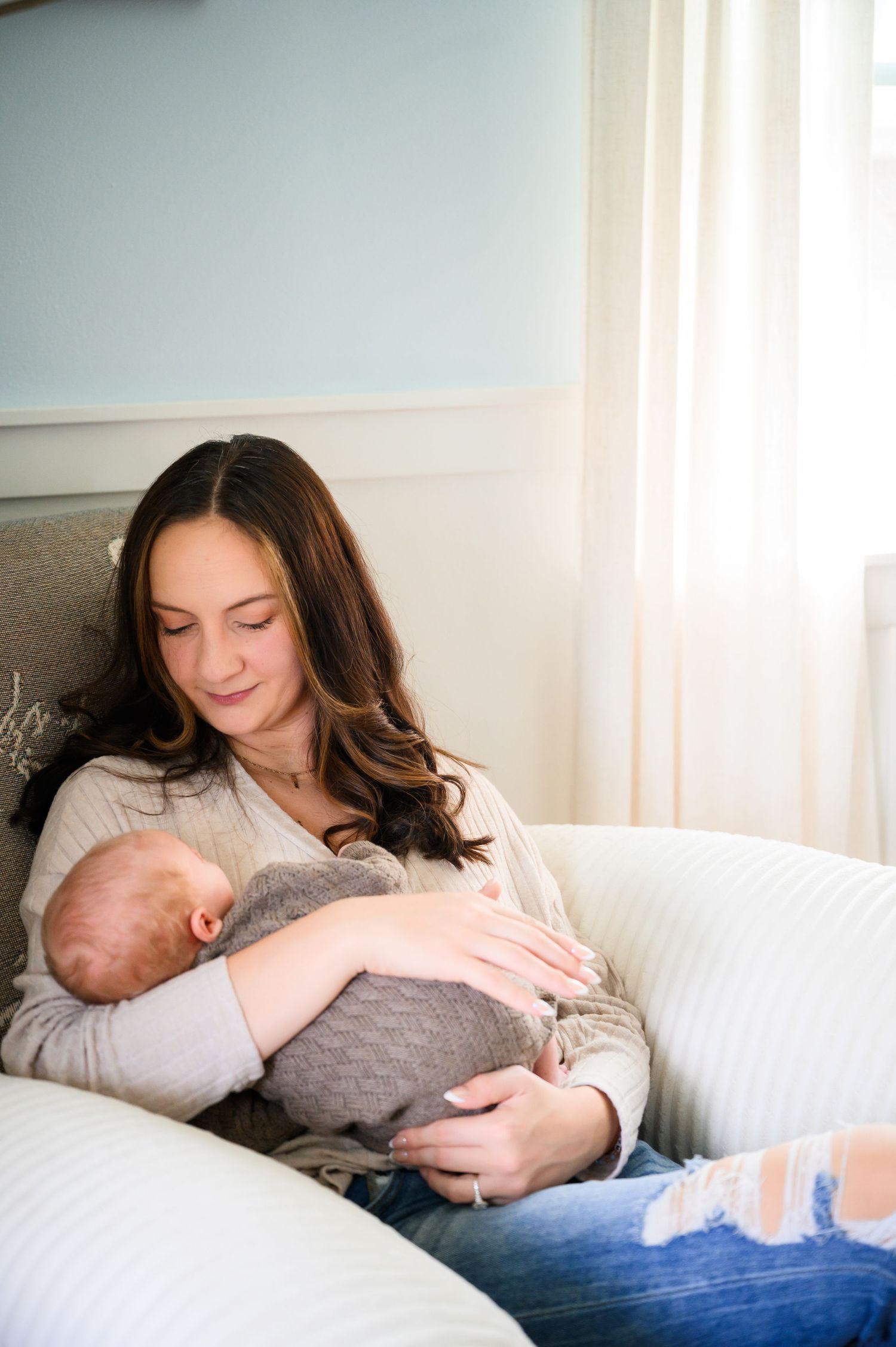 A young mother with long brown hair tenderly holds her newborn baby while sitting on a white sofa near a sunlit window.
