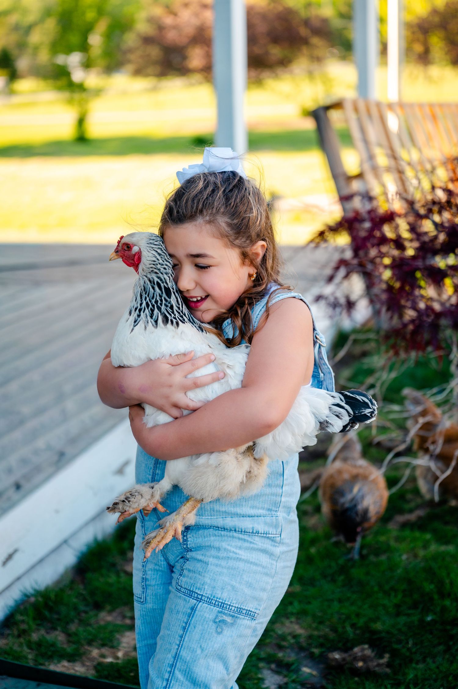 Young girl in denim overalls smiling while hugging a white chicken on a farm.