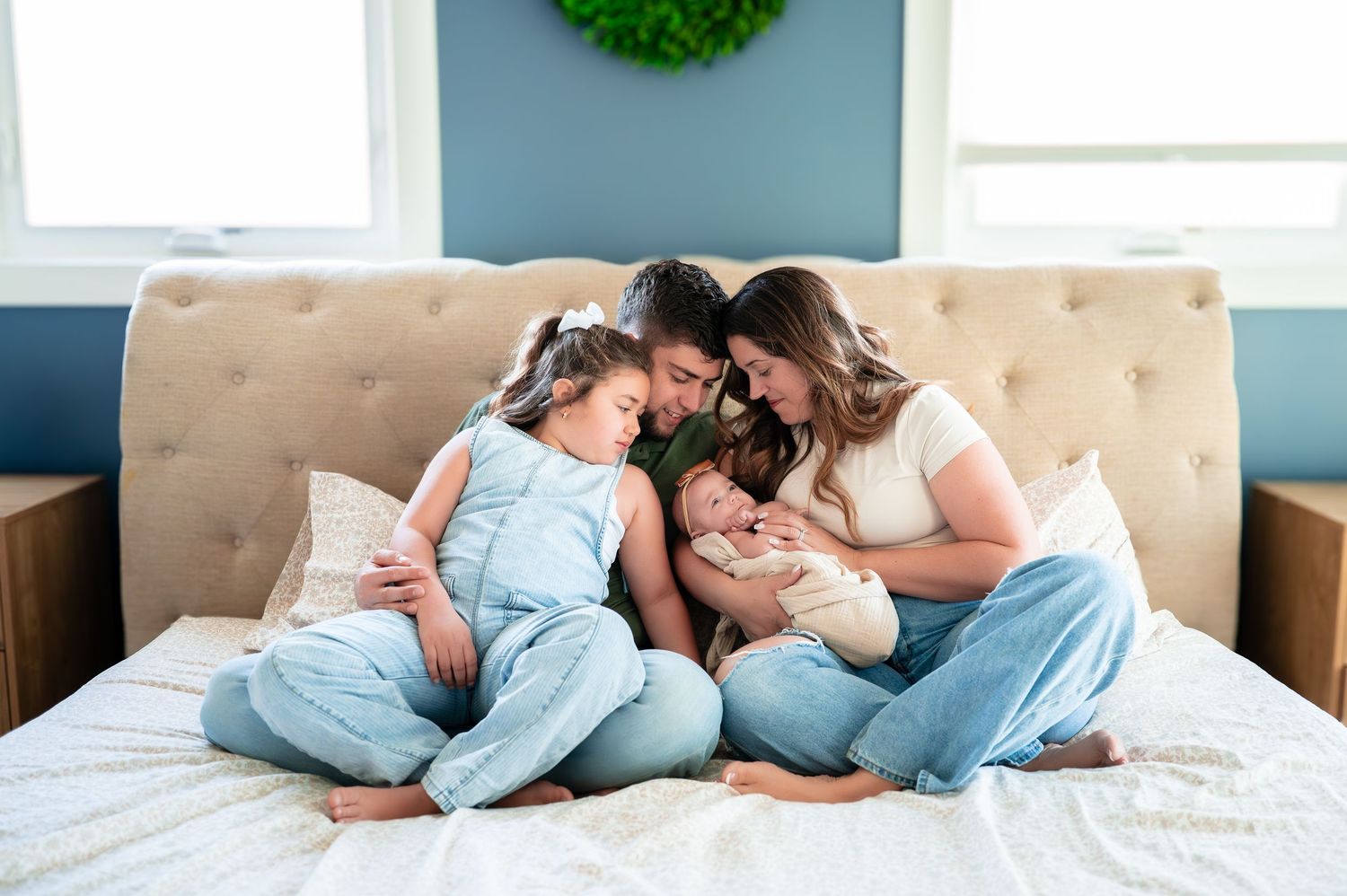 Family of four sitting on a bed, parents leaning in to admire their newborn baby while a young child cuddles close.