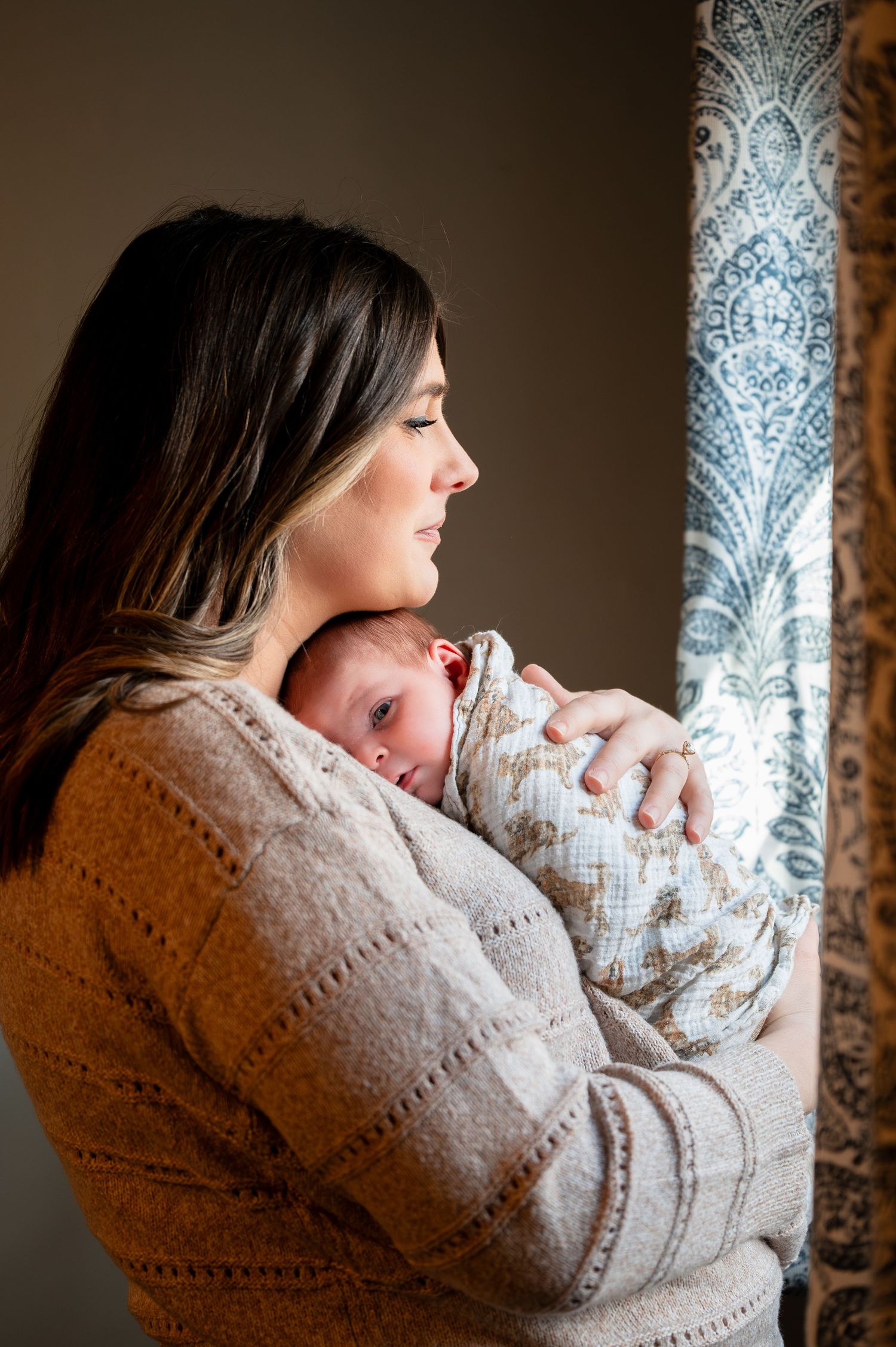 A mother tenderly holds her swaddled newborn baby near a window with floral curtains in warm indoor lighting.