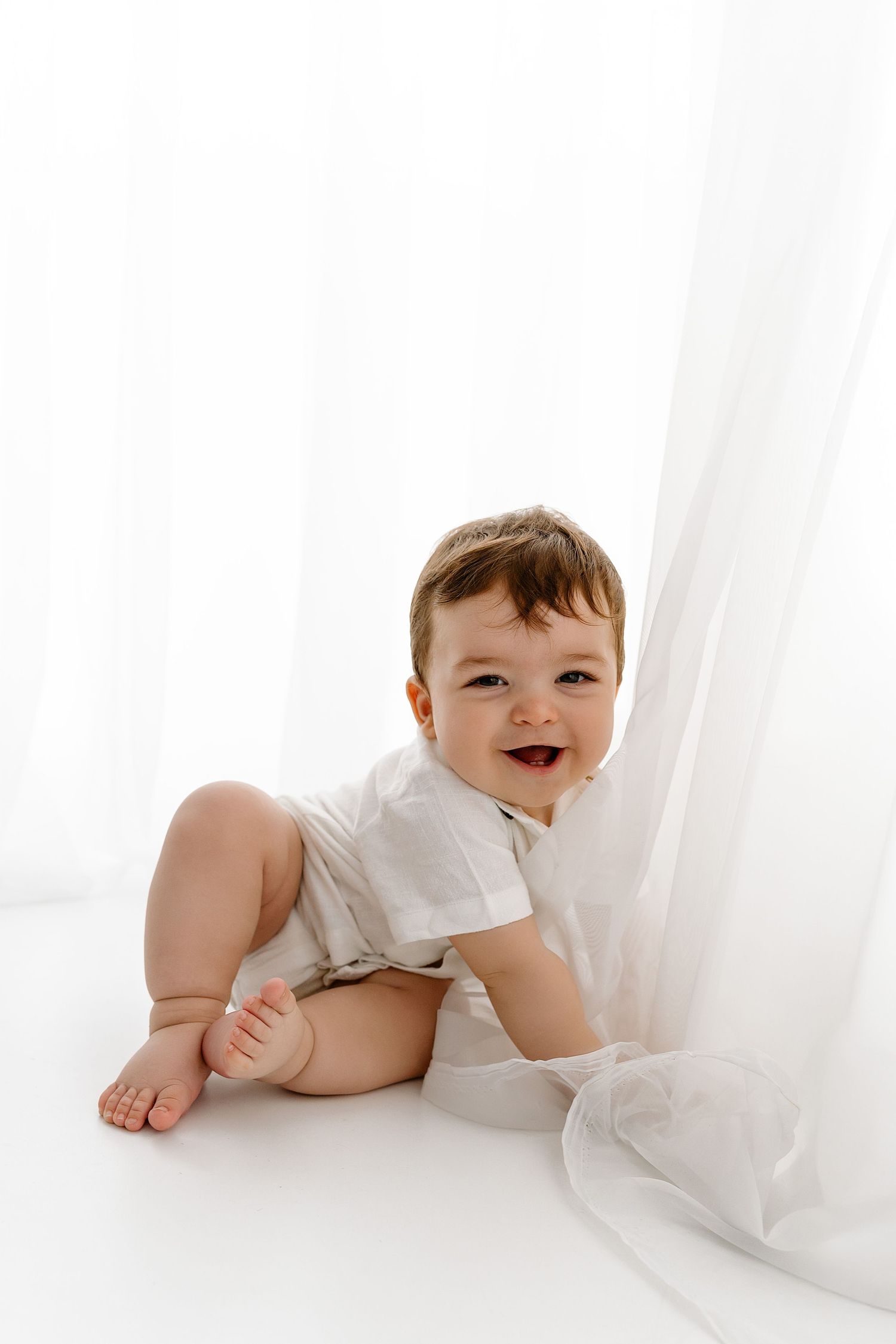 Small baby in white clothing sits on a white backdrop with sheer curtains, smiling brightly.