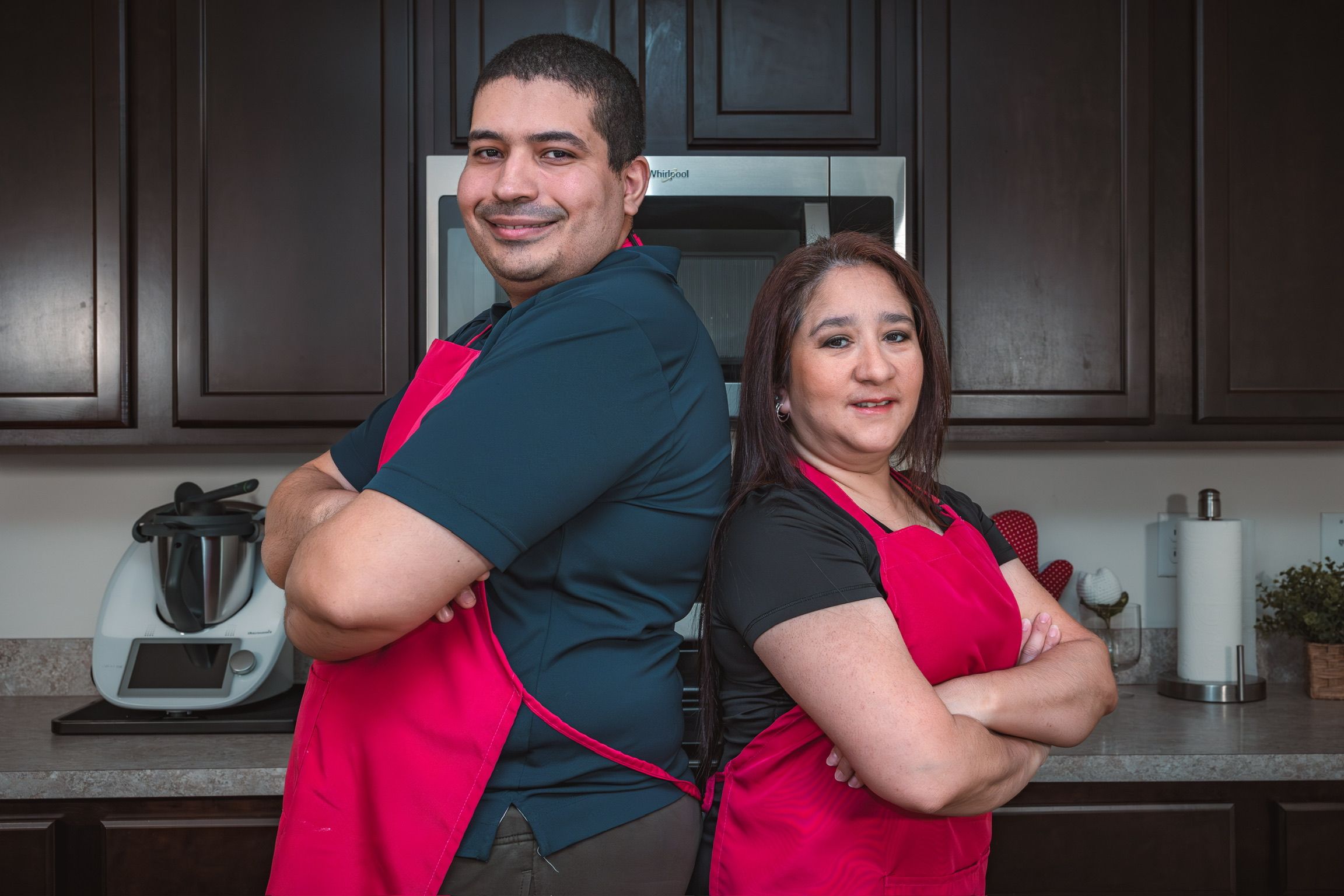 Two bakers wearing red aprons stand together in a modern kitchen setting.
