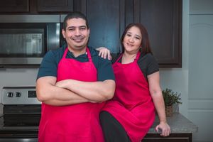 Bakers in red aprons pose in front of dark kitchen cabinets.