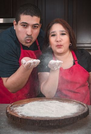 Bakers dusting powdered sugar over a dark wooden surface in a kitchen.