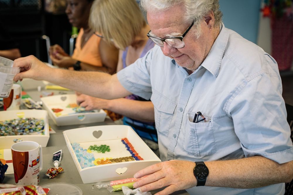 Senior in light blue shirt concentrates on creative art project at community workshop table.