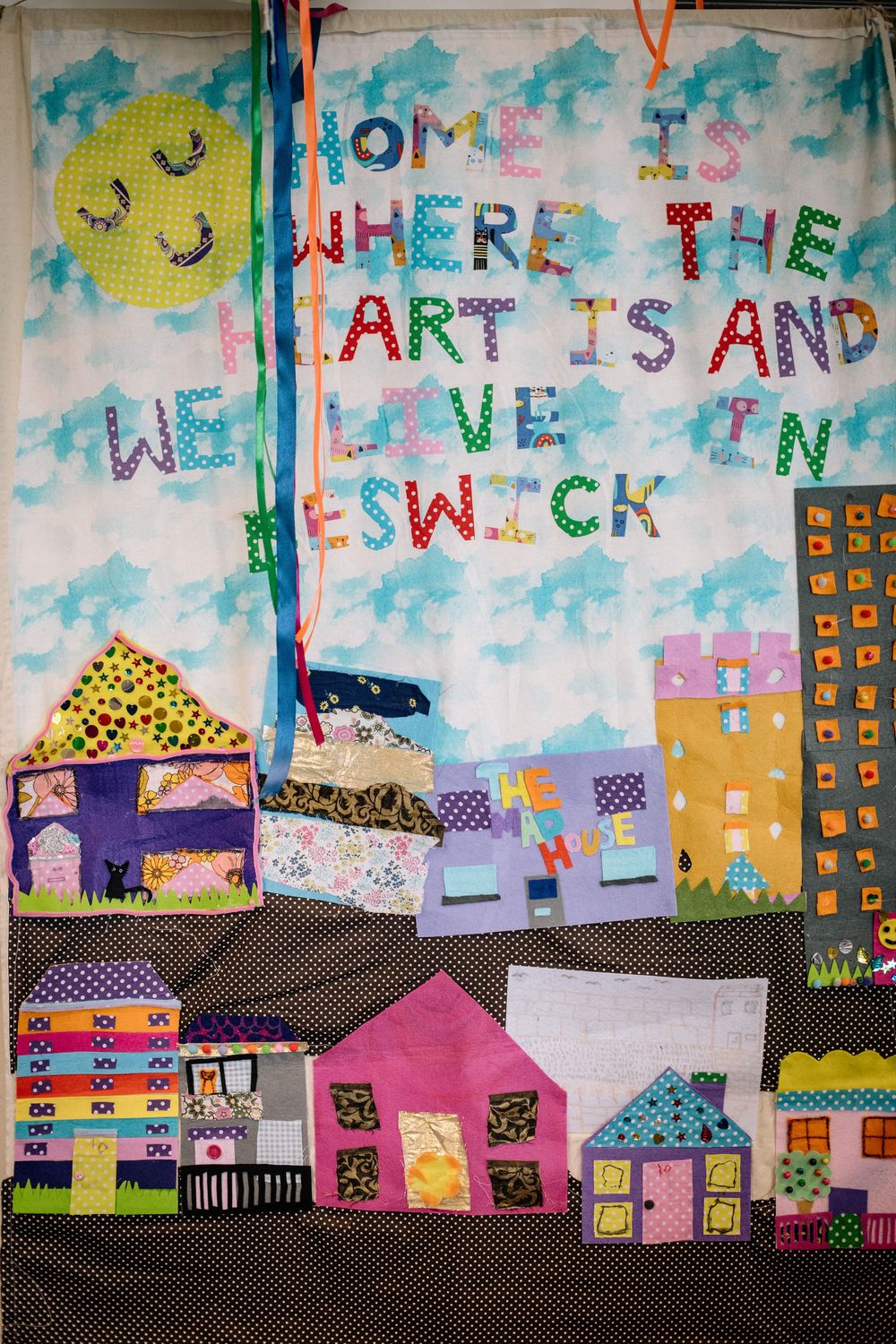 Colorful quilt display showing houses and text reading 'Home where the art is and we love Fenwick' against blue sky background.