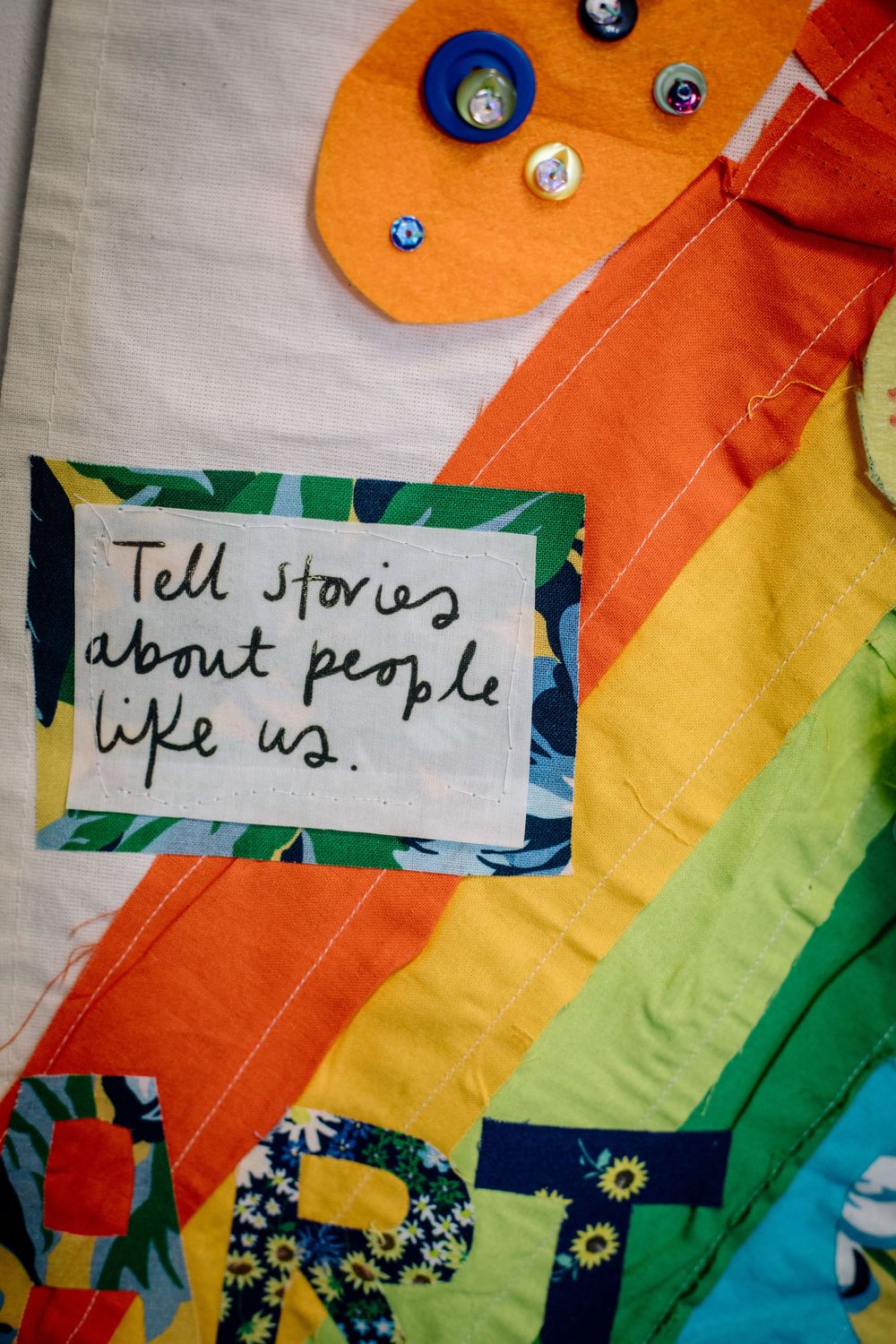A handwritten note reading 'Tell stories about people like us' placed on a colorful rainbow striped fabric background.