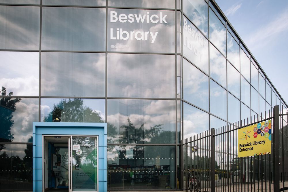 Modern glass library building exterior with reflective windows and metal framing against blue sky with clouds.