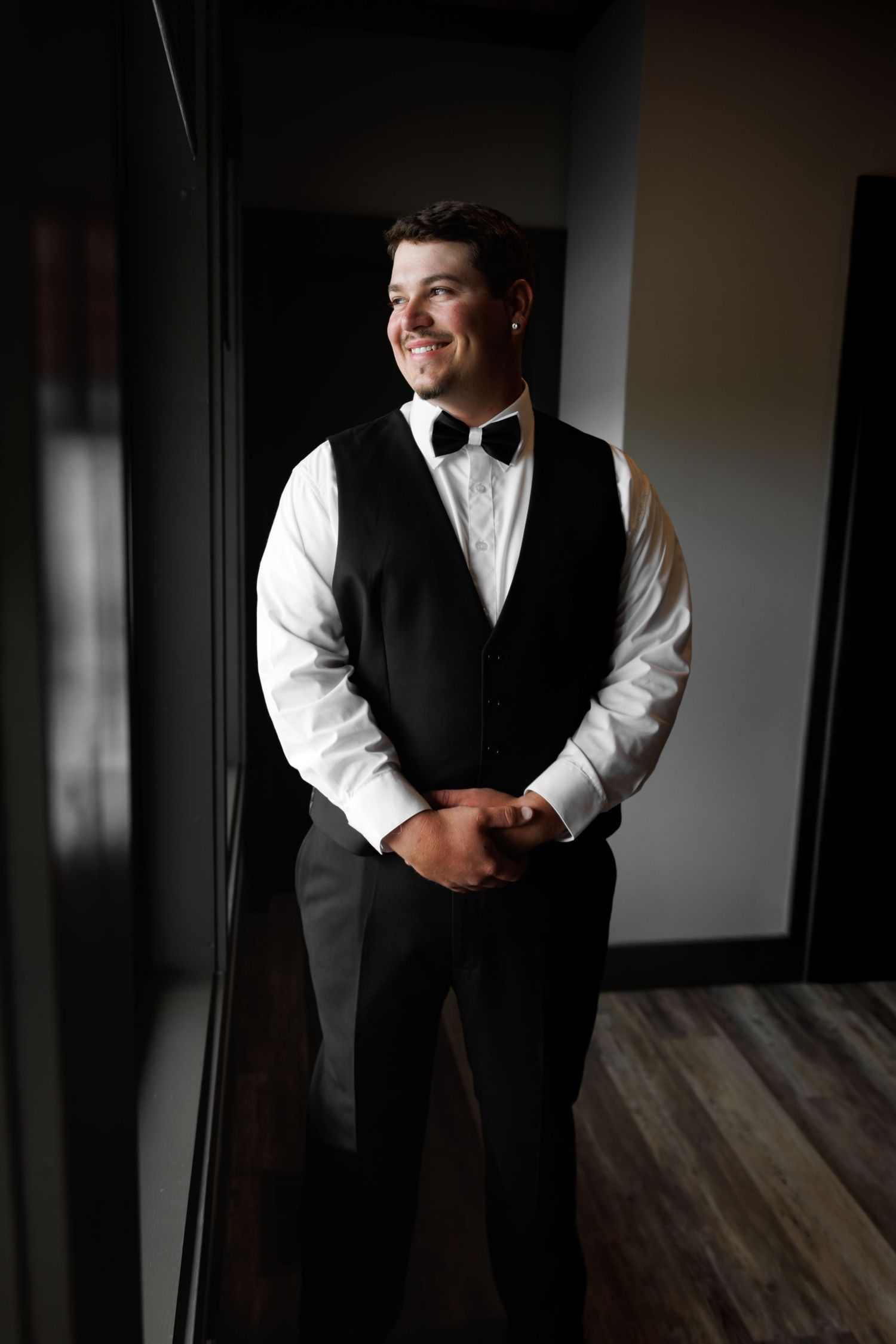 Smiling groom in black vest and bow tie standing by window in elegant wedding attire.