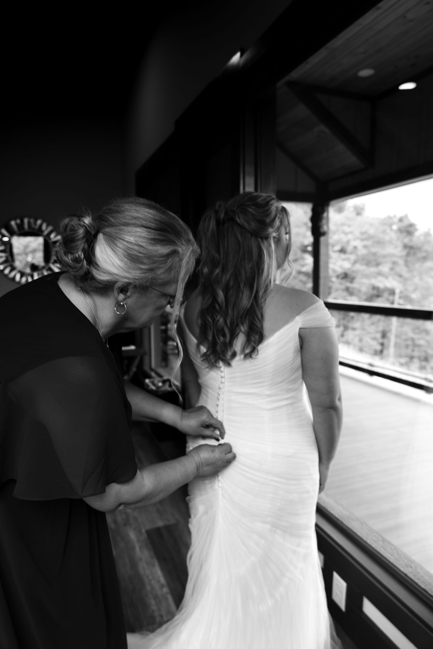 Black and white photo of a woman helping a bride button her wedding dress near a window.