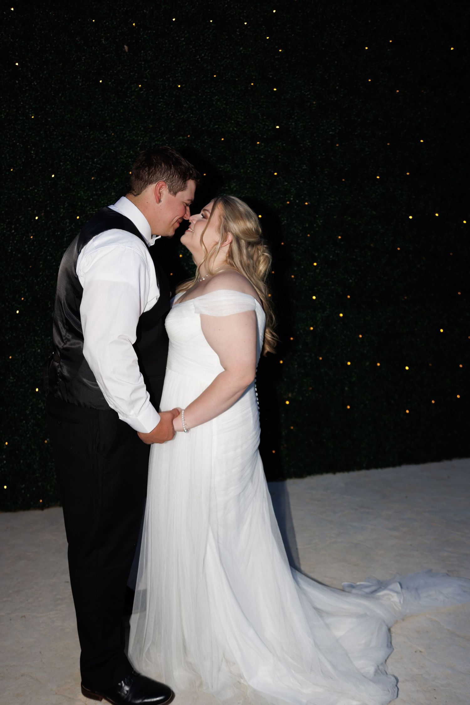 Bride and groom share a romantic almost-kiss during their first dance against a glittering greenery backdrop.