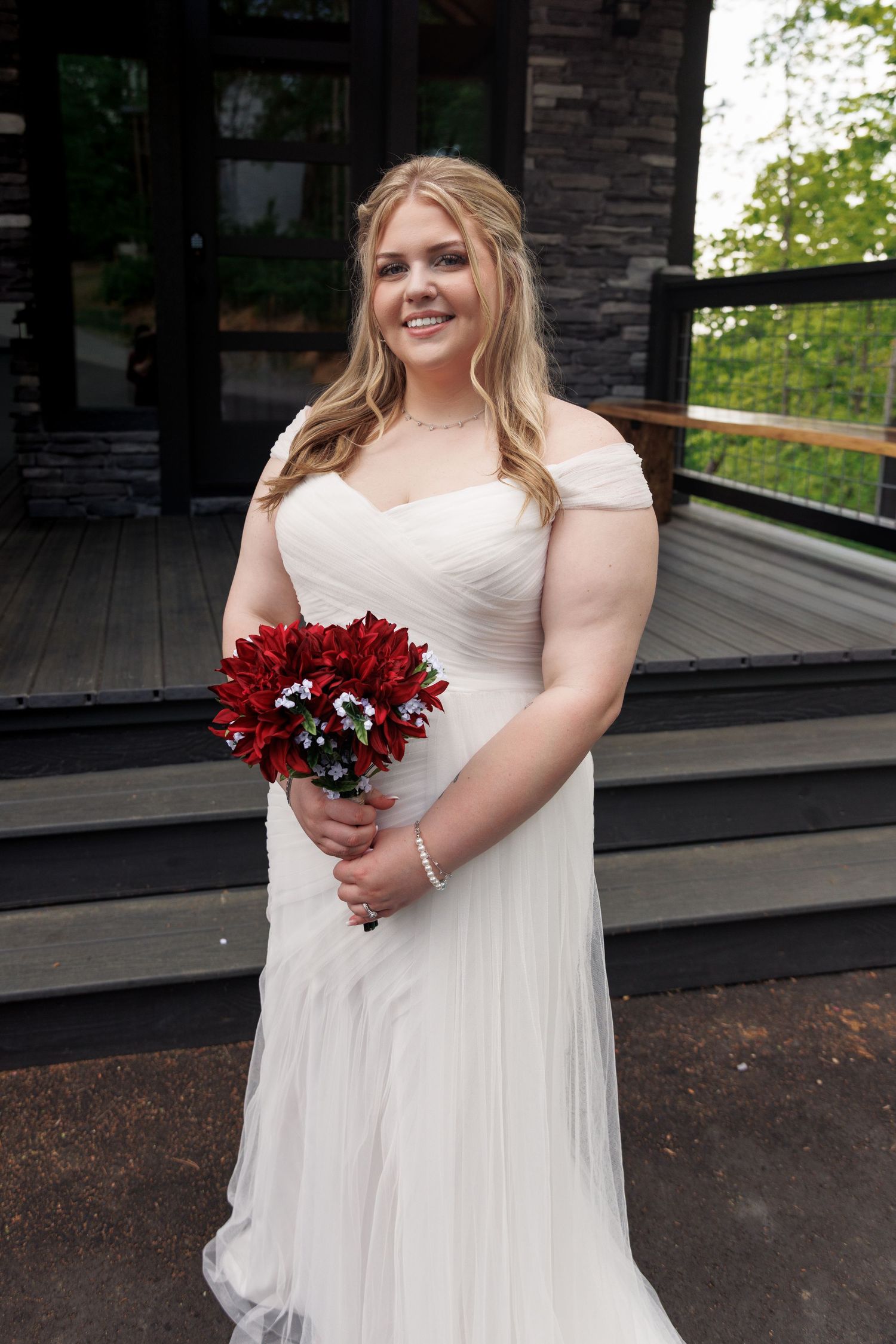 Smiling bride in white off-shoulder gown holding red floral bouquet on wooden deck surrounded by greenery.