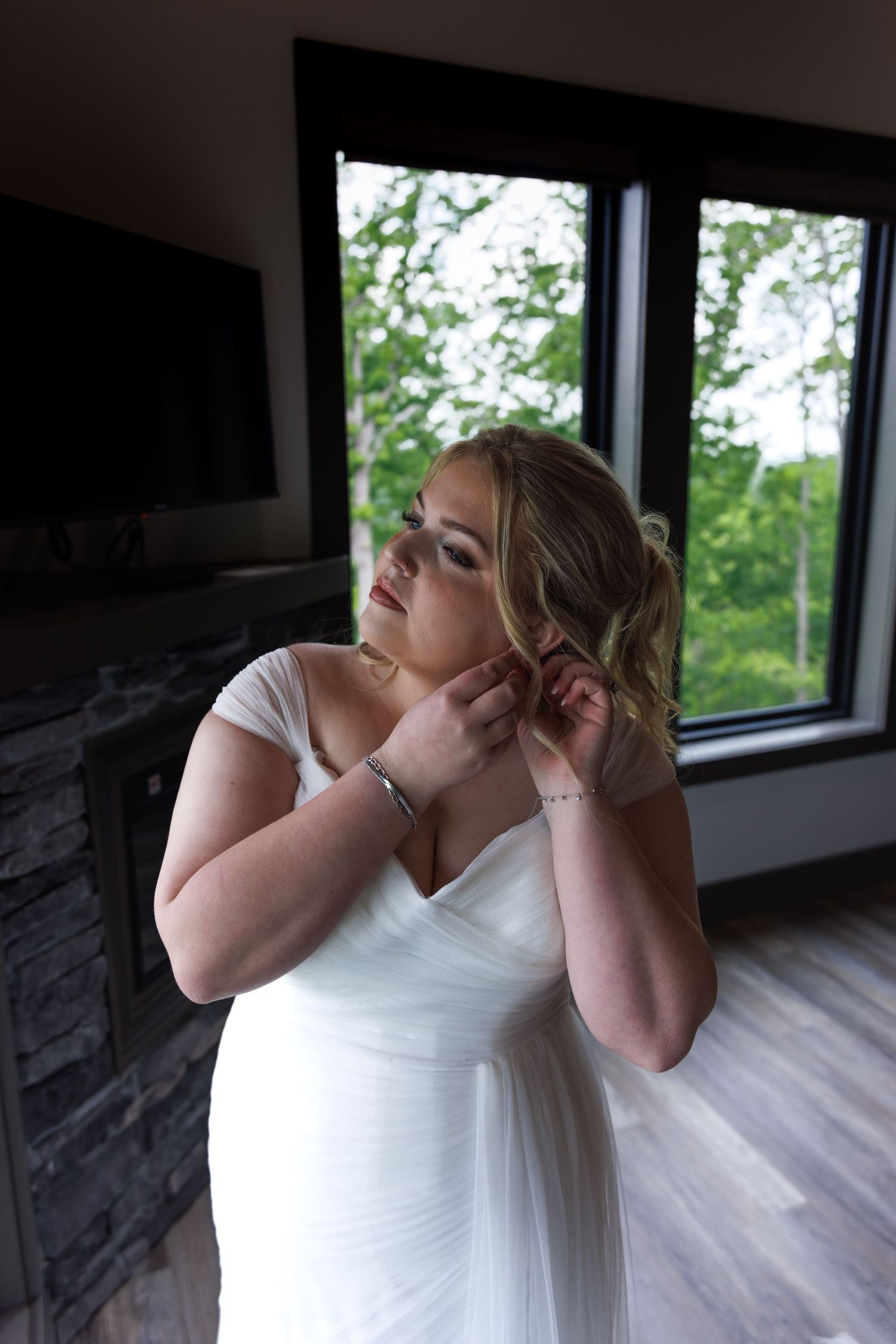 Elegant bride in white off-shoulder gown adjusting earrings by a window on her wedding day.