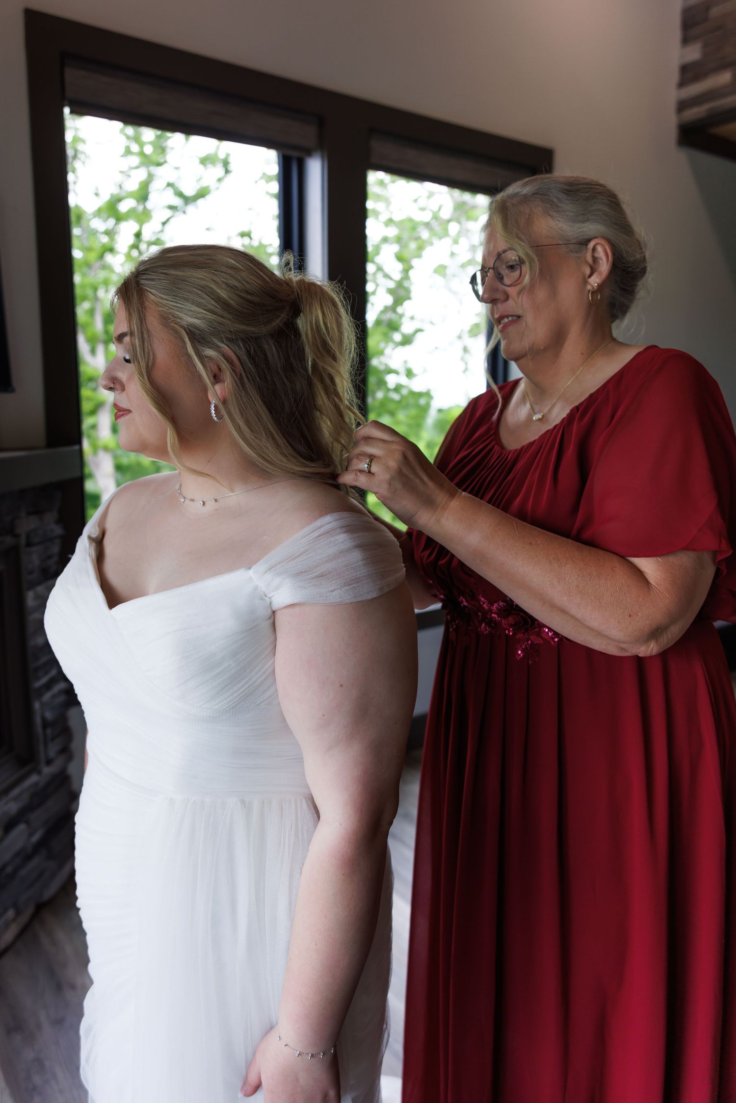 Woman in red dress helping a bride with her white wedding gown near a window.