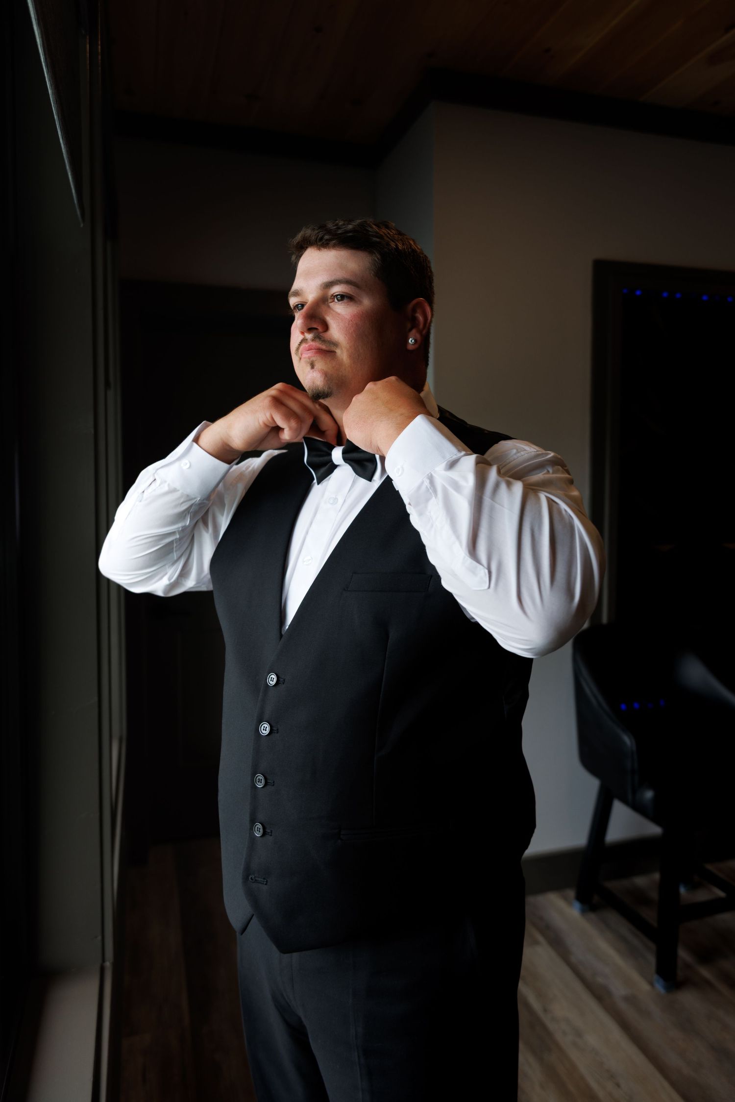 Groom adjusting bow tie wearing black vest and white dress shirt before wedding ceremony.
