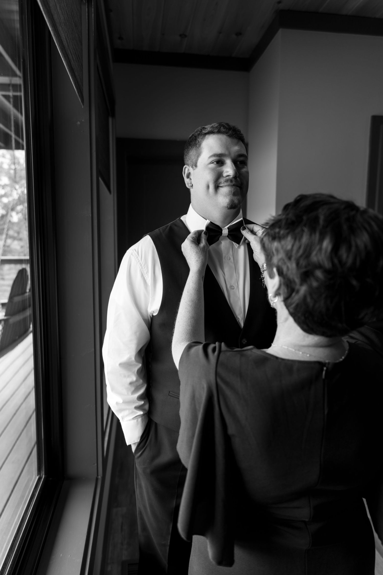Groom smiling as someone adjusts his bow tie near a window in black and white wedding photo.