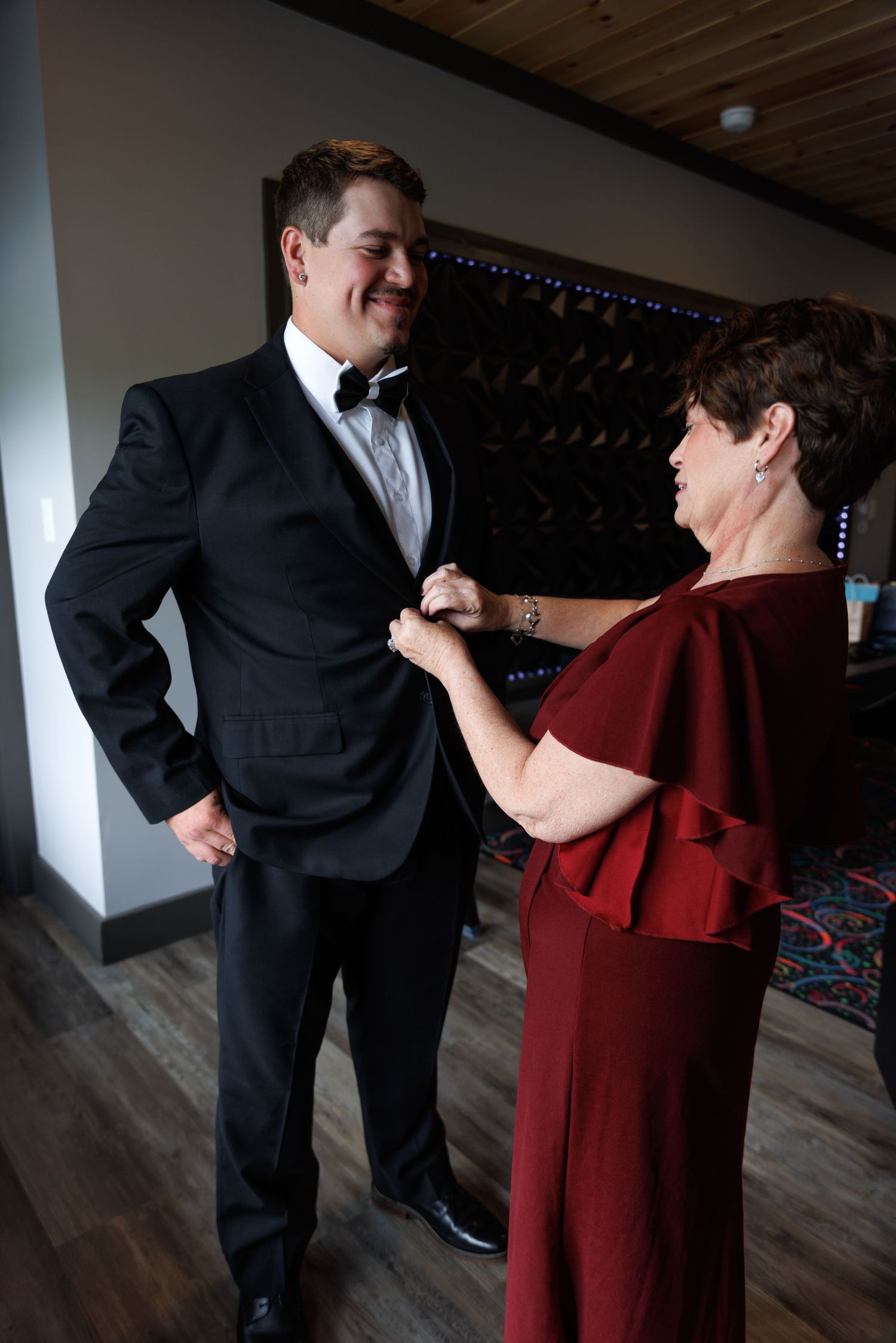 Smiling groom in black tuxedo gets boutonniere pinned by woman in red dress before wedding.