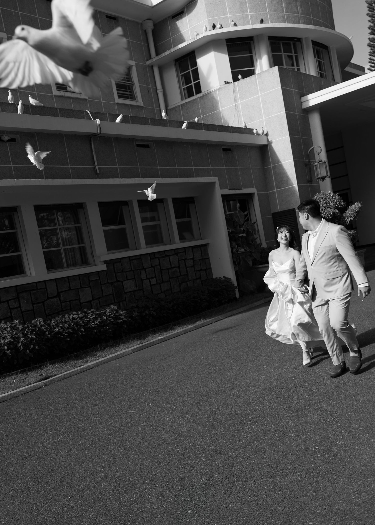 Black and white photo of a couple in wedding attire walking outside a building with doves flying overhead.
