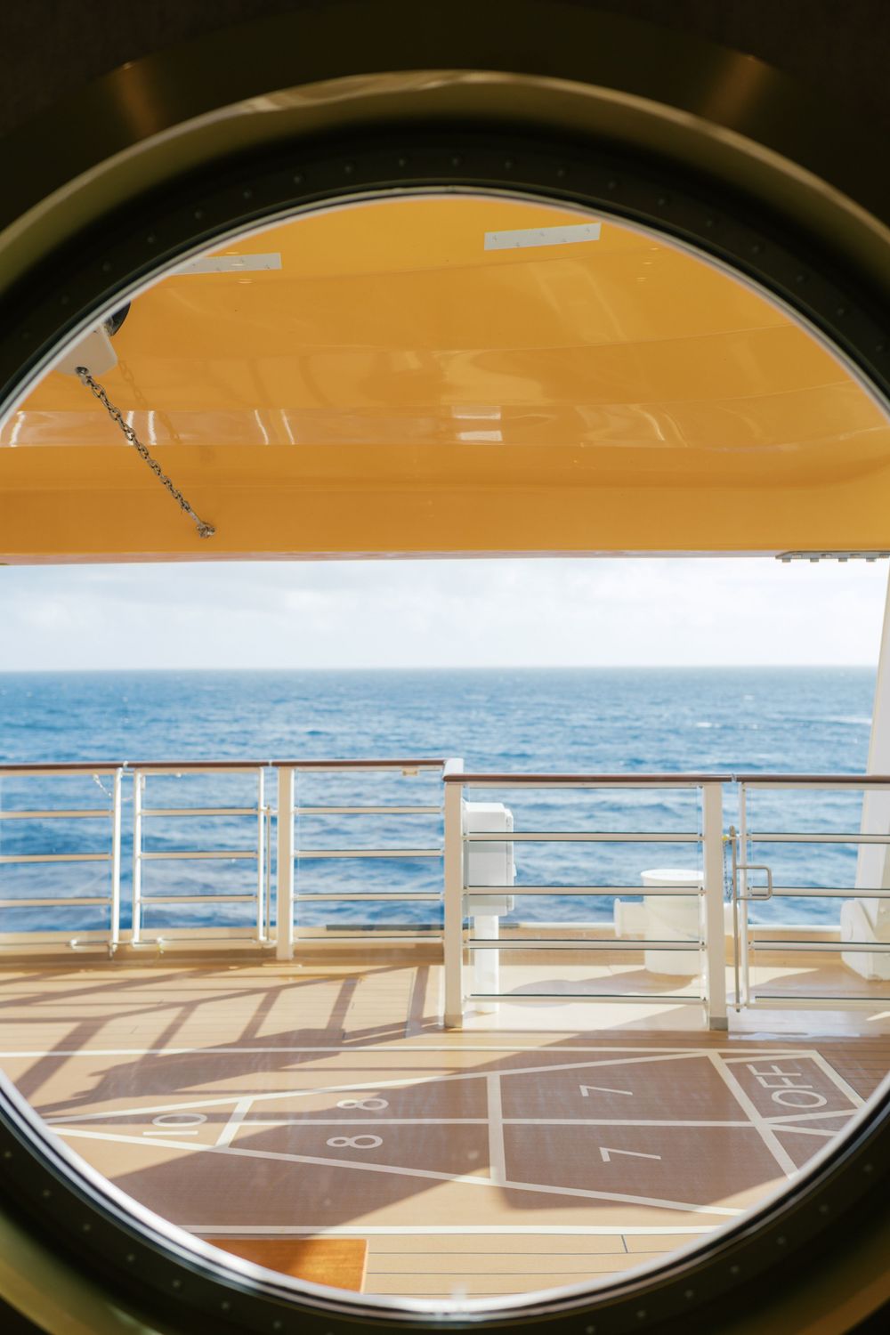 View through a cruise ship porthole of the ocean deck with shuffleboard court, railing, and calm blue sea beyond.