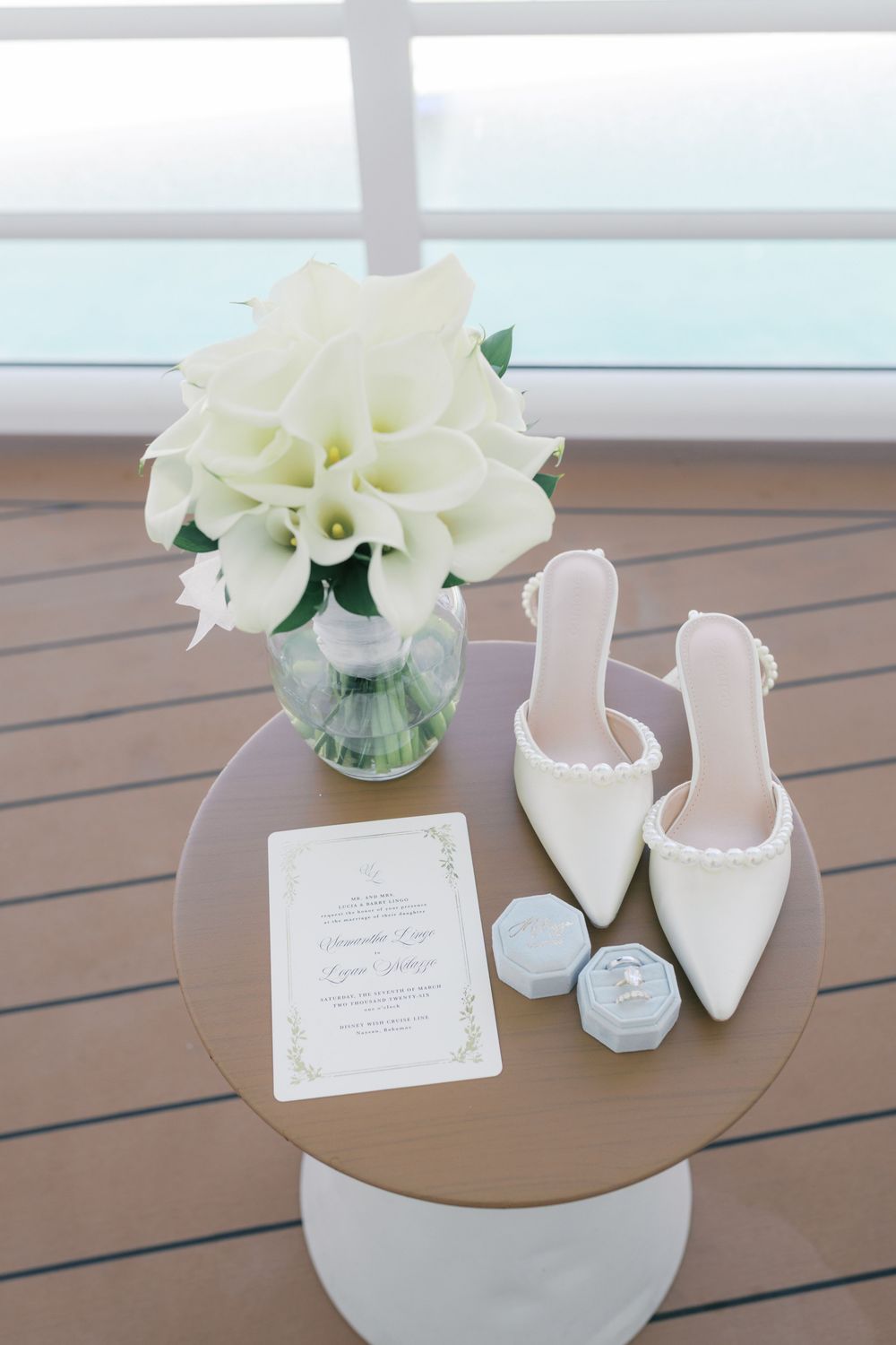 White bridal heels, calla lily bouquet, ring box, and wedding invitation styled on a round table by a window.