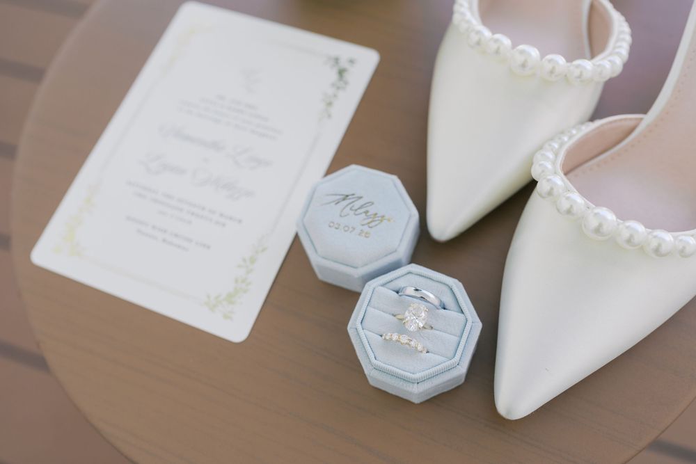 Wedding flat lay with pearl heels, ring box with diamond rings, and elegant invitation on a wooden surface.