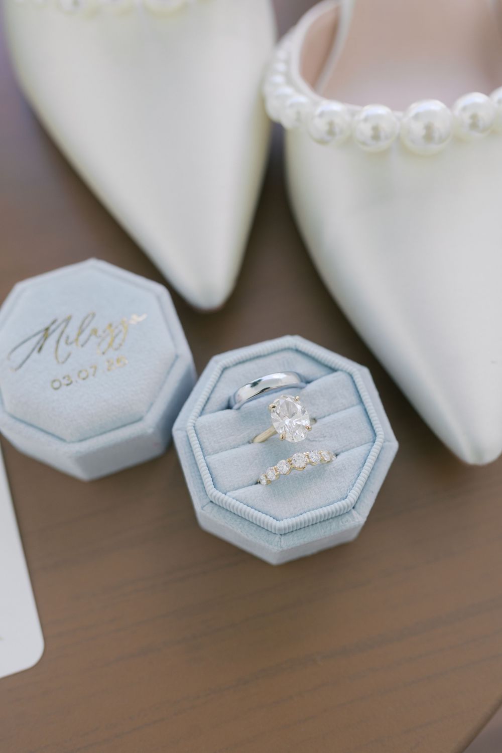 Diamond engagement ring and wedding band in a blue velvet ring box beside pearl-trimmed bridal heels.