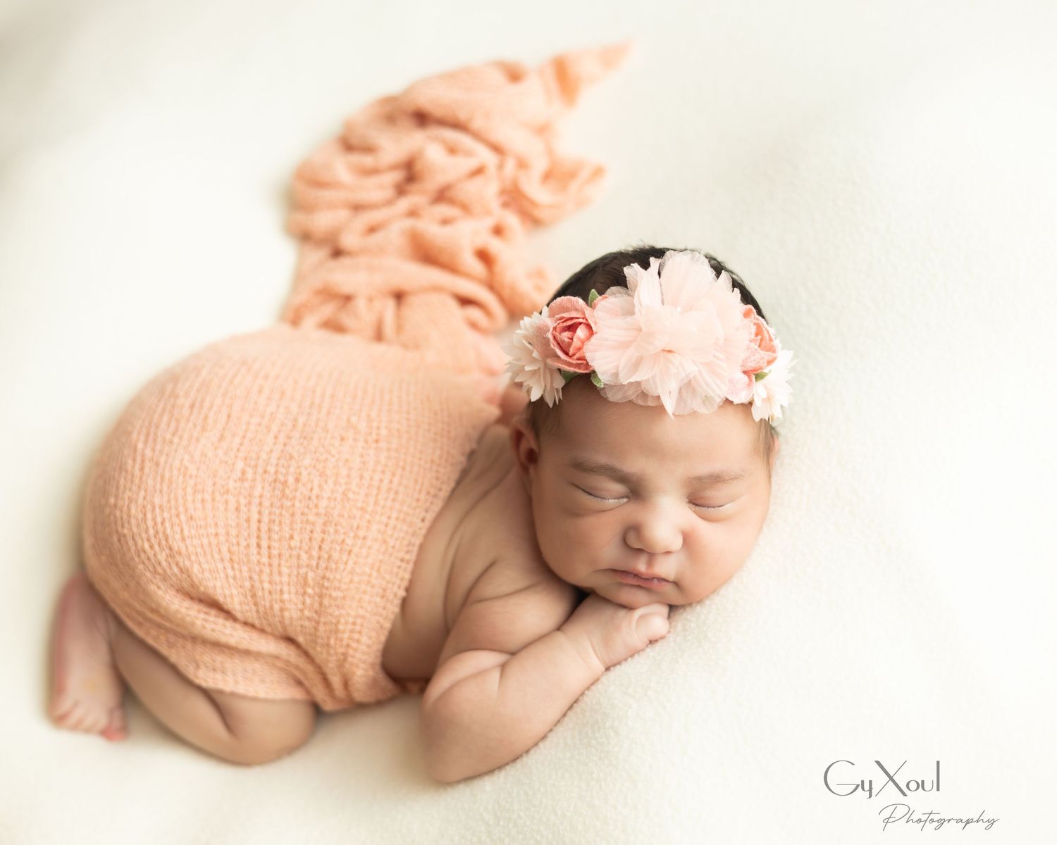 Sleeping newborn baby girl in peach wrap and floral headband posed on white fur for professional photography.