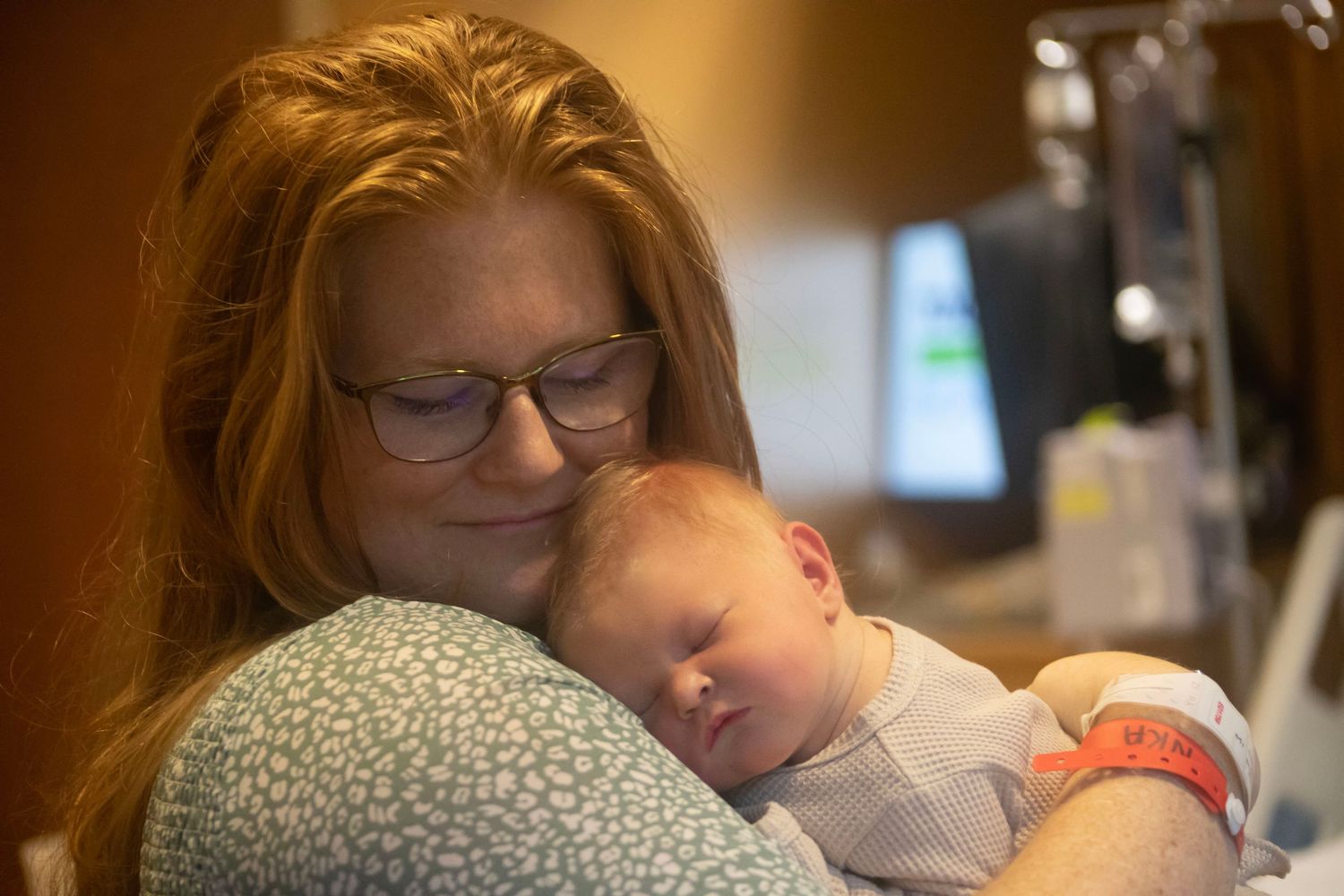 A red-haired woman wearing glasses tenderly holds a sleeping newborn baby in a hospital room.